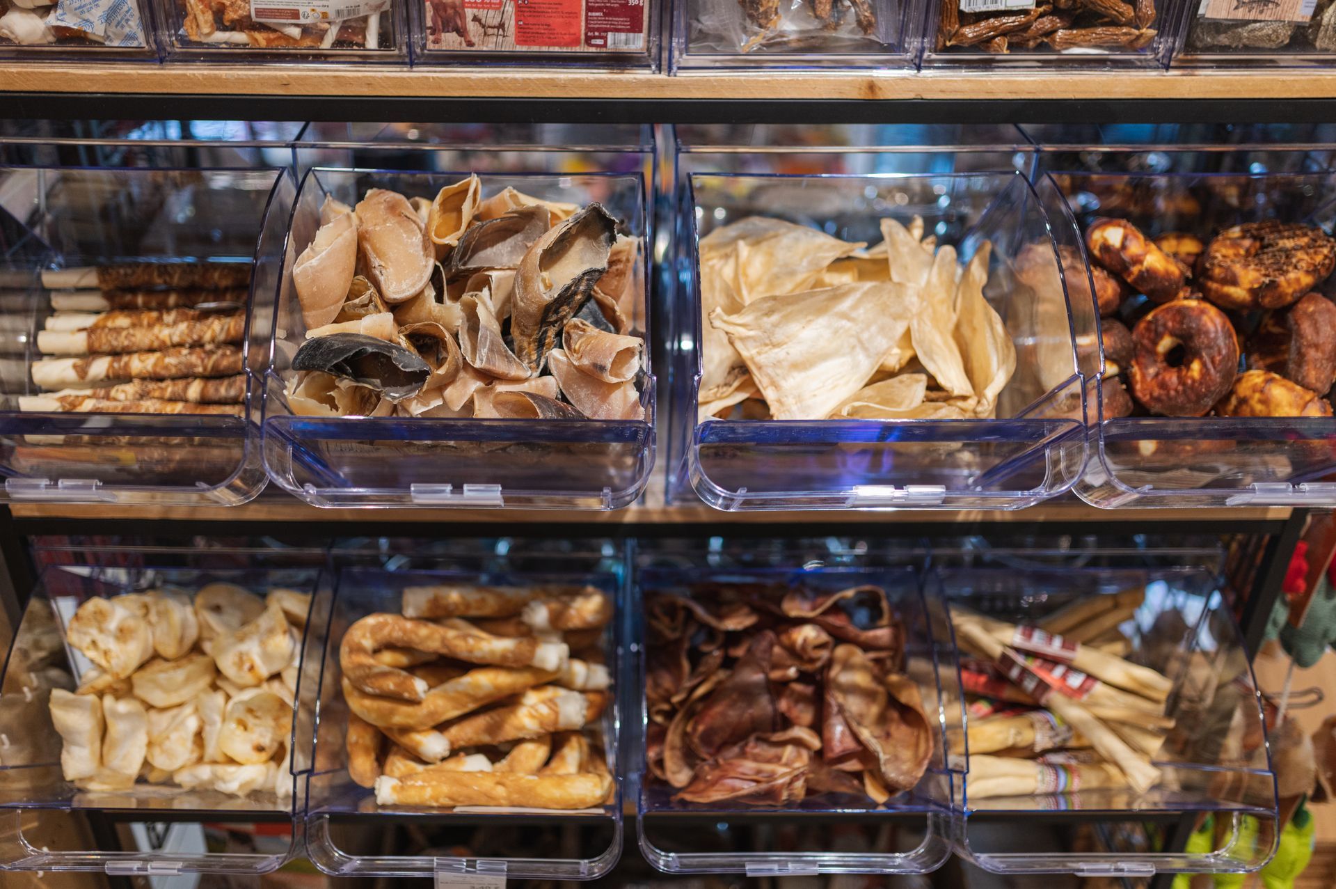 Des friandises pour chiens dans des boîtes transparentes sur une étagère en bois, de formes et de textures variées.