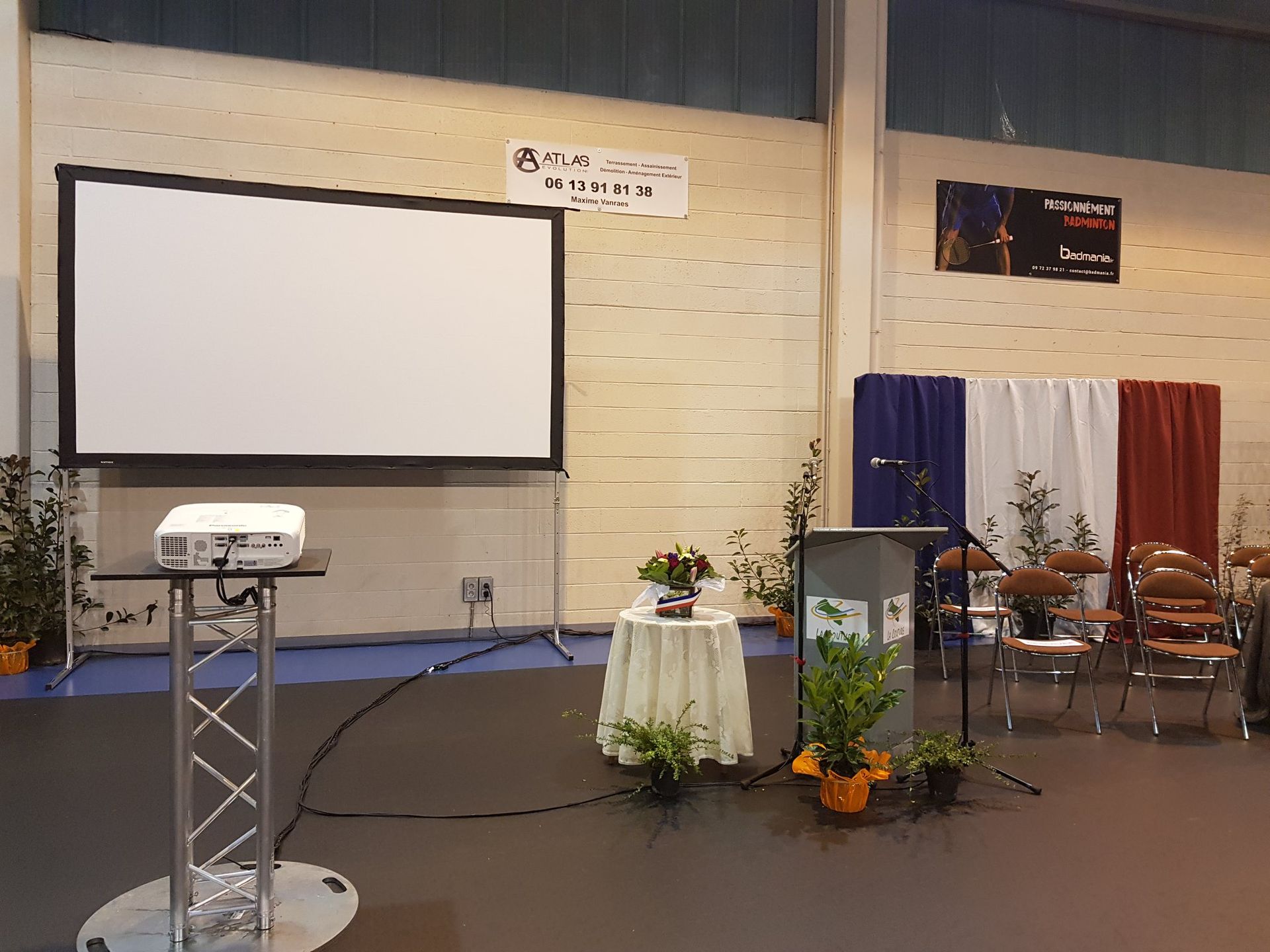 Scène de conférence vide avec écran de projection, podium, chaises et rideaux rouges, blancs et bleus près des fleurs.