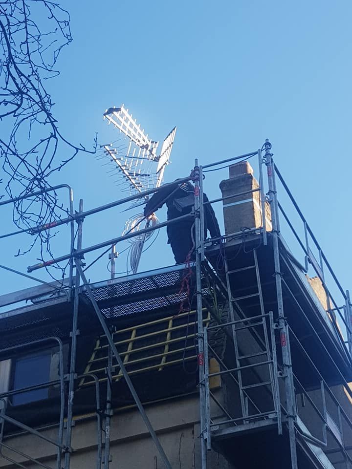 Un ouvrier sur un échafaudage à côté du toit d'un bâtiment, avec une antenne parabolique sur fond de ciel bleu.