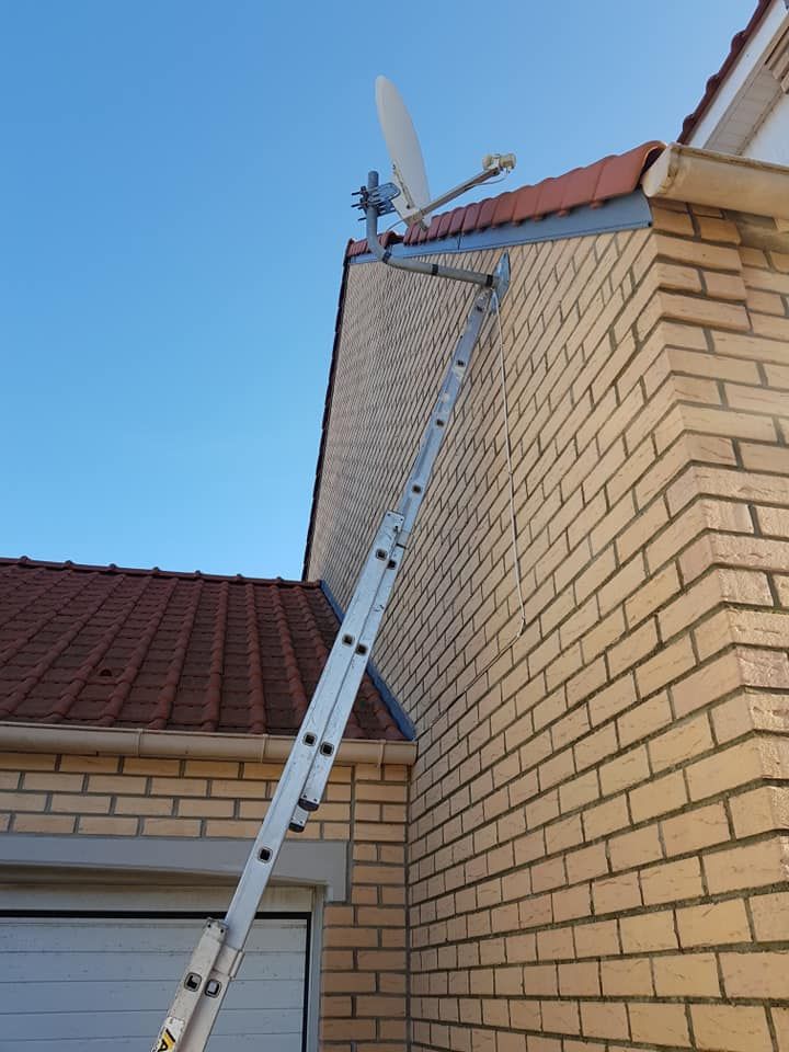 Une échelle est appuyée contre le toit en briques d'une maison, à côté d'une cheminée, sous un ciel bleu limpide.