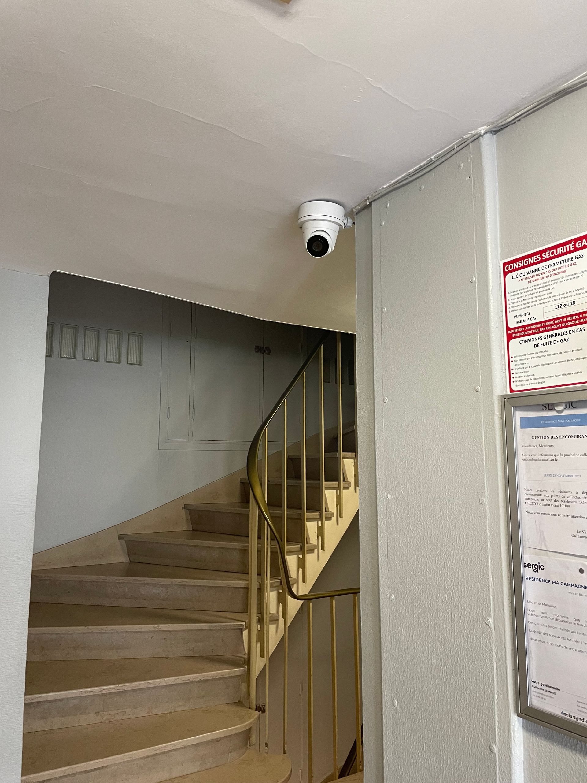 Escalier intérieur avec marches beiges, rampe en métal et caméra de sécurité au plafond.
