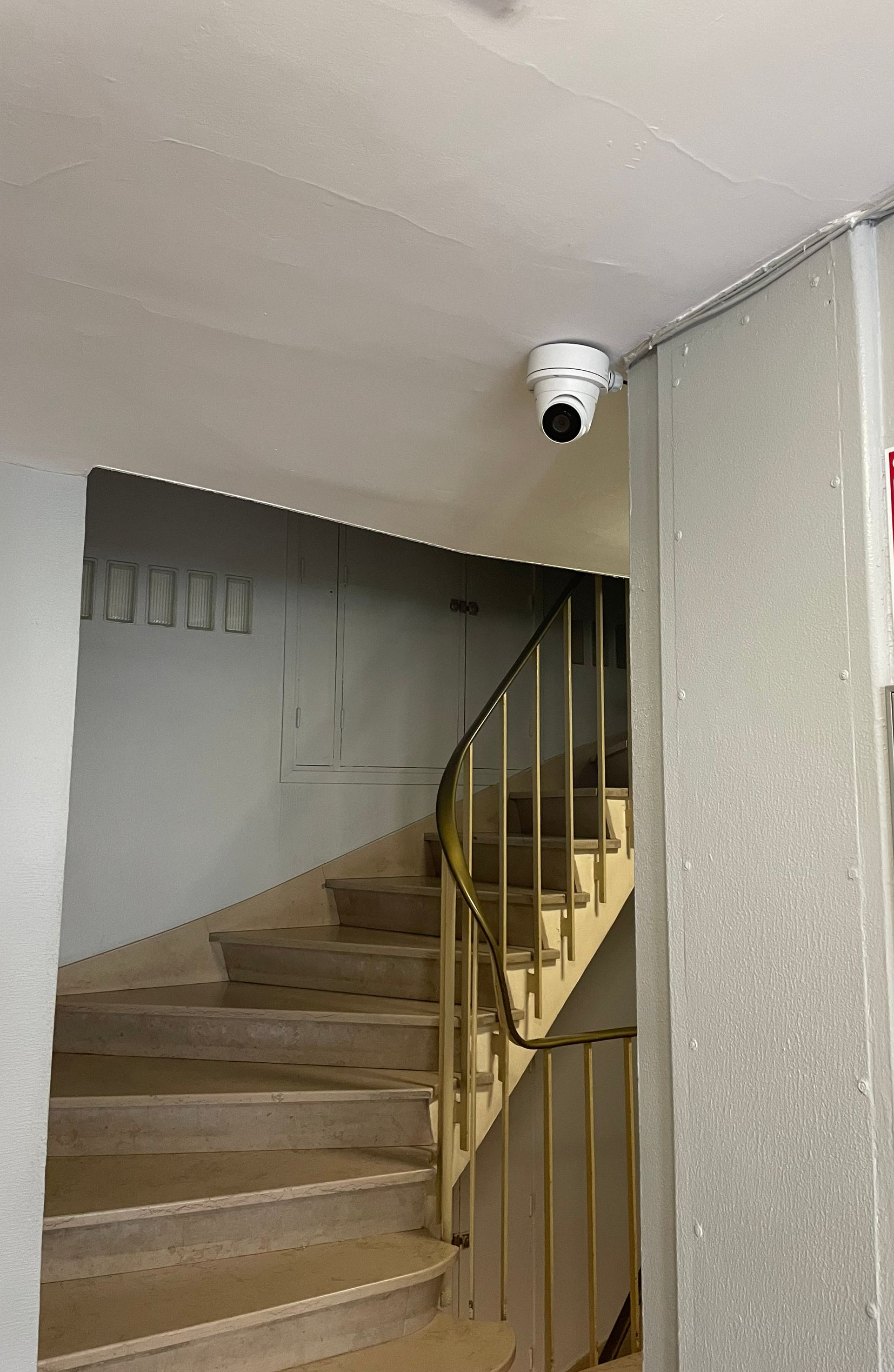 Escalier intérieur avec marches beiges, rampe en métal et caméra de sécurité au plafond.