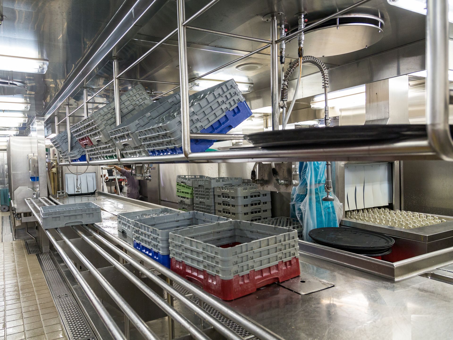 Station de lavage de vaisselle commerciale en acier inoxydable avec des caisses en plastique grises et bleues empilées sur des tapis roulants.