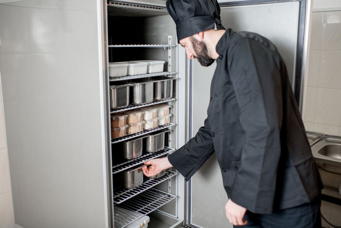 Un chef en uniforme noir plonge la main dans un grand réfrigérateur industriel rempli de conteneurs de stockage métalliques.