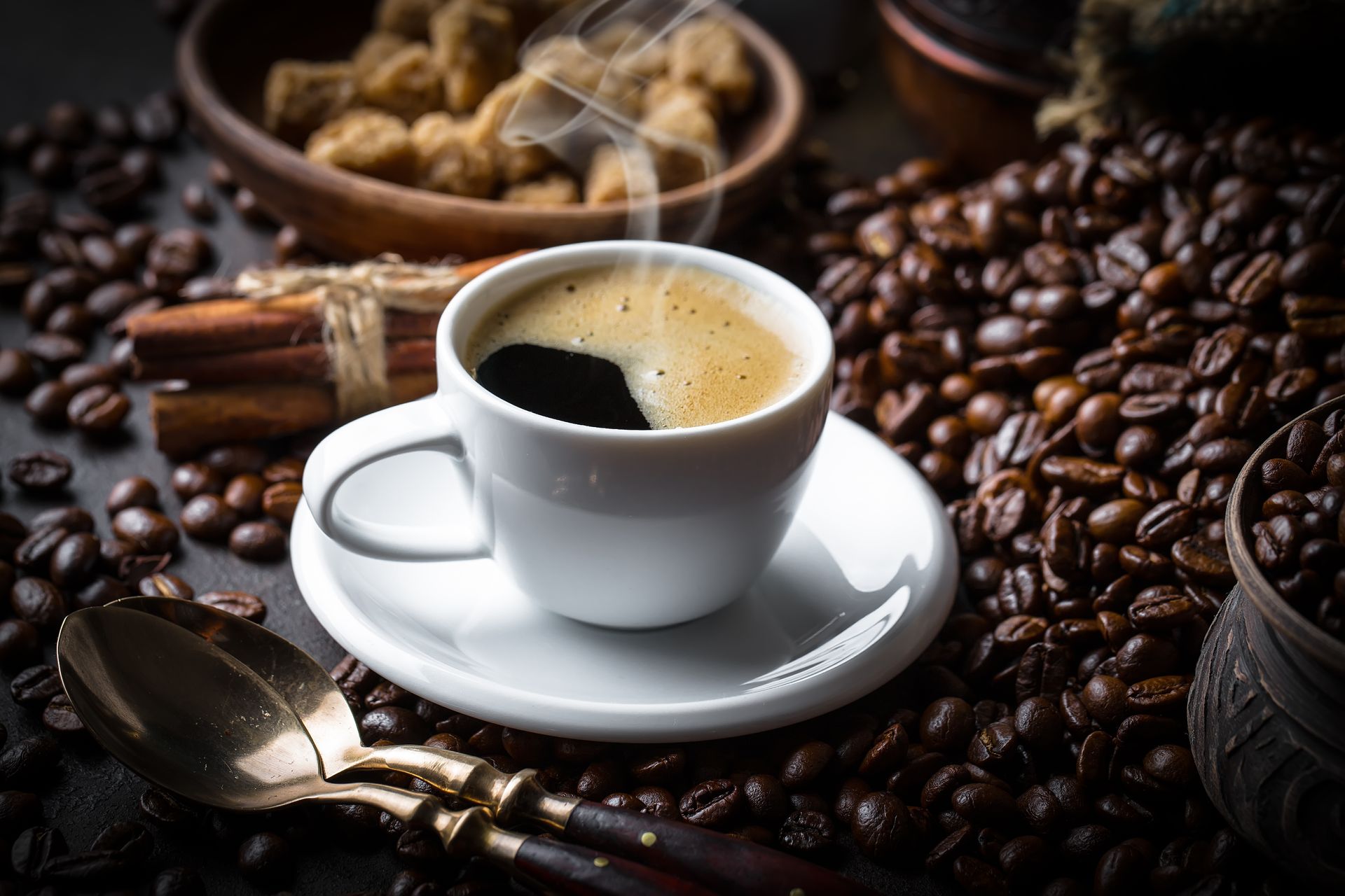 Une tasse blanche de café noir fumant posée sur une soucoupe, entourée de grains de café torréfiés, d'une cuillère et de bâtonnets de cannelle.