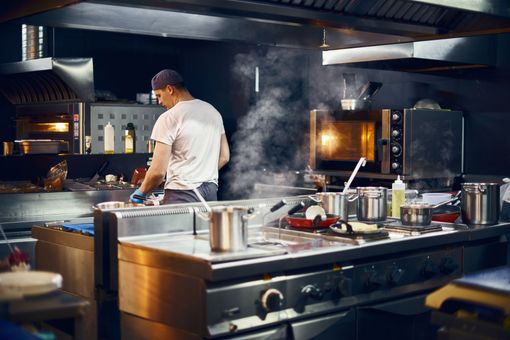 Un chef en t-shirt blanc et casquette travaille devant un fourneau de cuisine professionnelle, d'où s'échappe de la vapeur des casseroles.