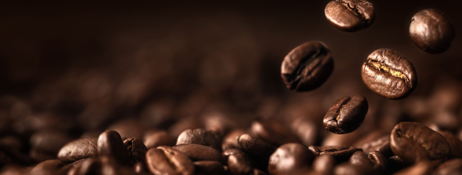 Grains de café torréfiés, dont plusieurs sont suspendus en l'air au-dessus d'un tas de grains foncés et texturés sur fond brun.