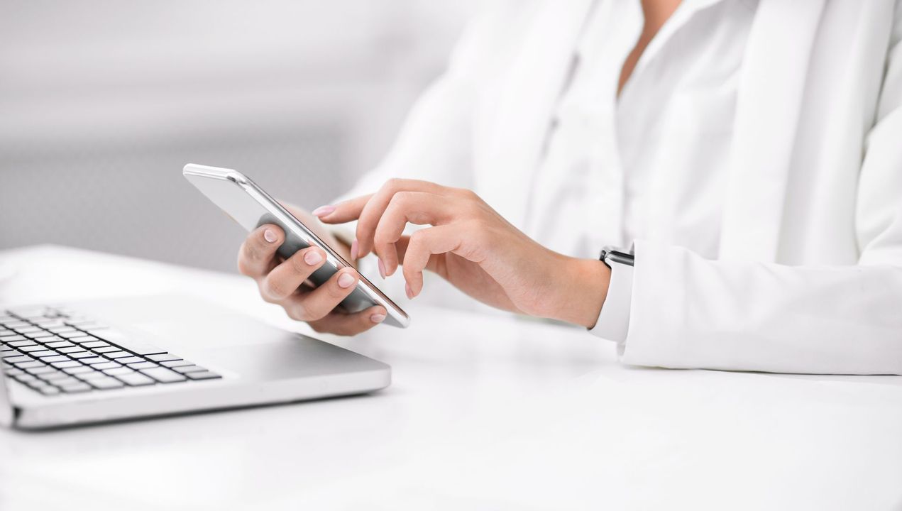 Une personne en costume blanc est assise à un bureau et utilise un smartphone, le clavier d'un ordinateur portable étant partiellement visible.