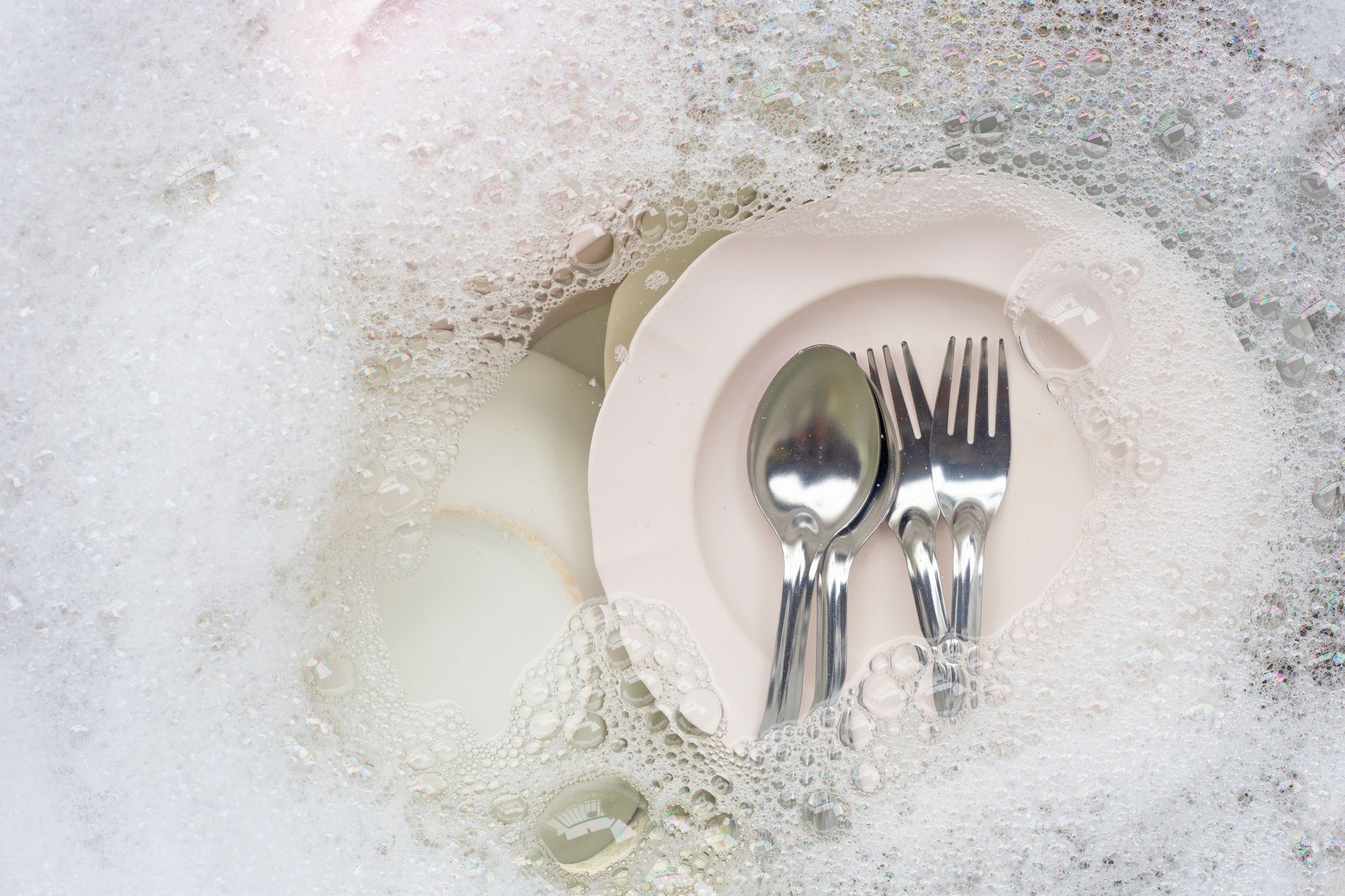 Une simple assiette avec une cuillère et une fourchette posées dessus, plongée dans un évier rempli d'eau savonneuse et de bulles.