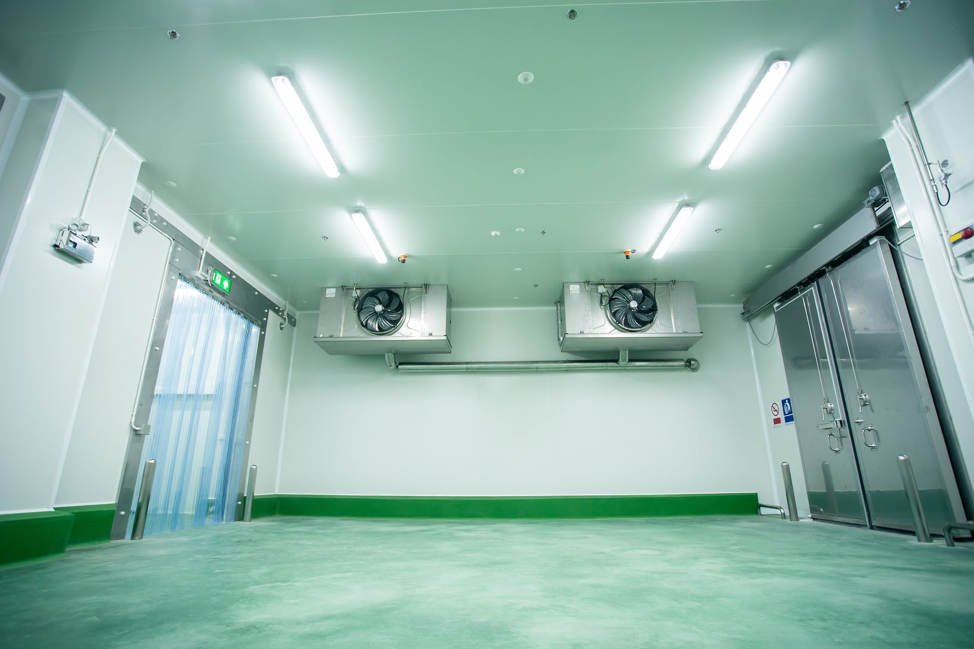 Une chambre froide industrielle blanche et propre, équipée d'un éclairage au plafond, de deux ventilateurs de refroidissement, d'une porte à lanières en plastique et de portes en acier.