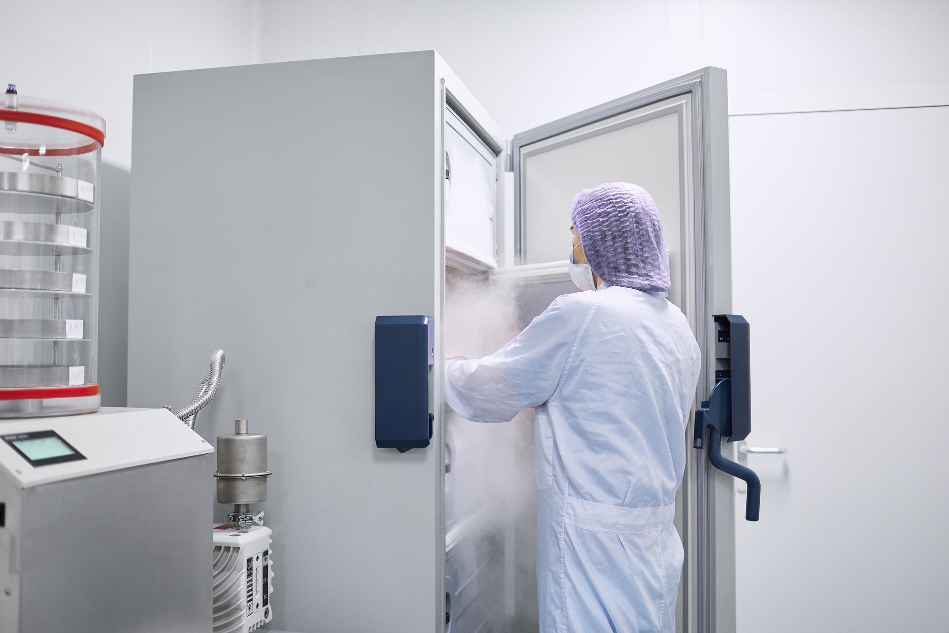 Une personne portant un équipement de protection ouvre un congélateur de laboratoire, provoquant l'échappement d'un nuage d'air froid ou de vapeur dans une salle blanche.