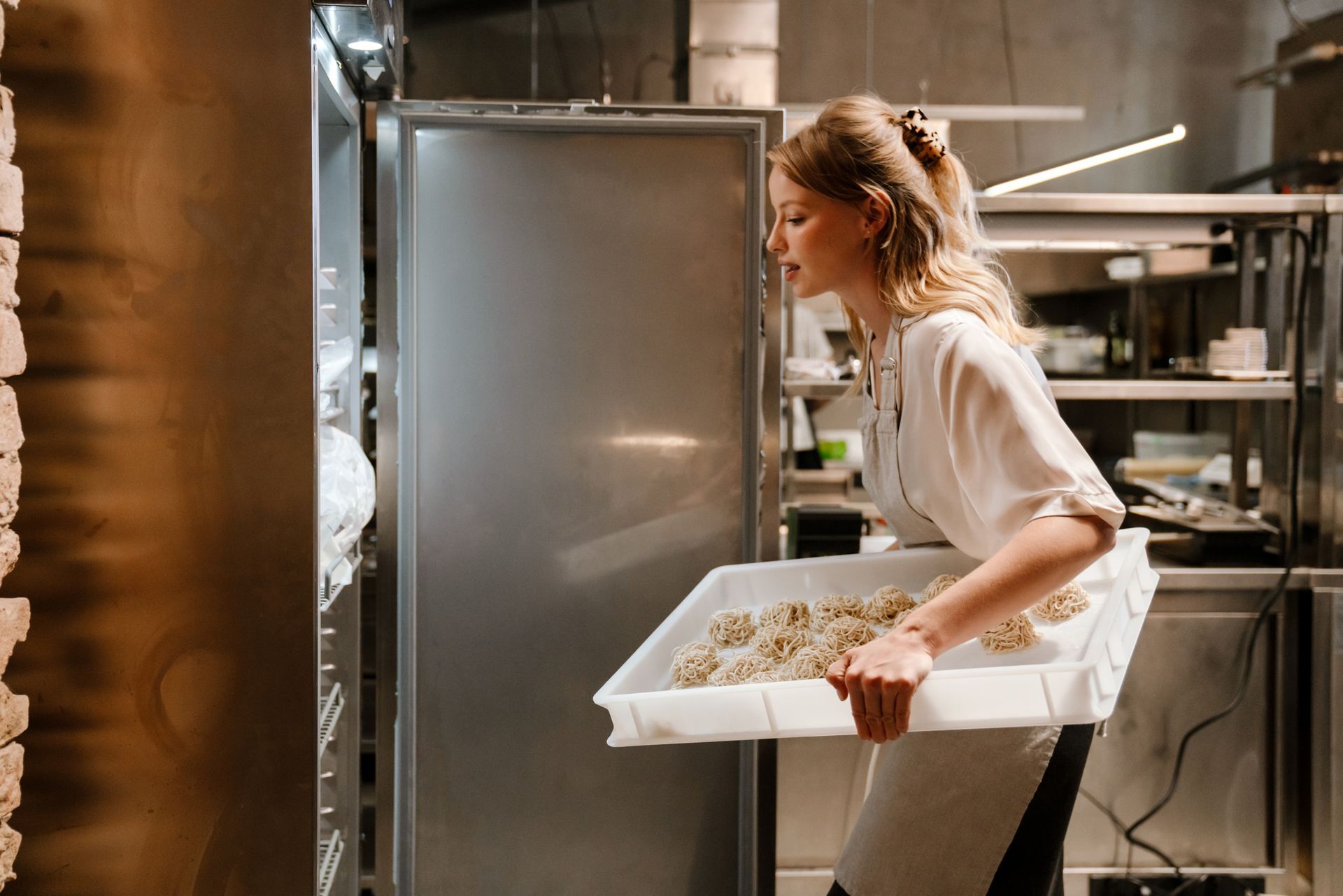 Une personne portant un tablier de cuisine place un plateau de viennoiseries crues dans un grand réfrigérateur industriel en acier inoxydable.
