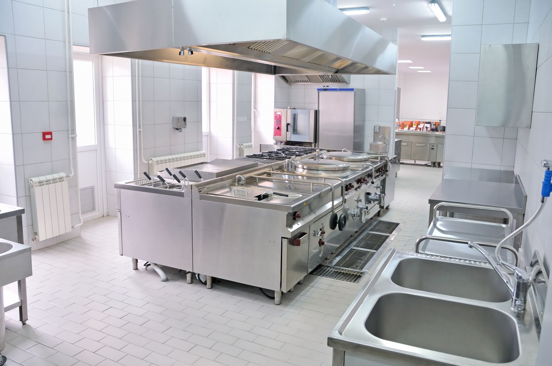 Une cuisine professionnelle et lumineuse avec des appareils de cuisson en acier inoxydable, une grande hotte aspirante et un double évier.