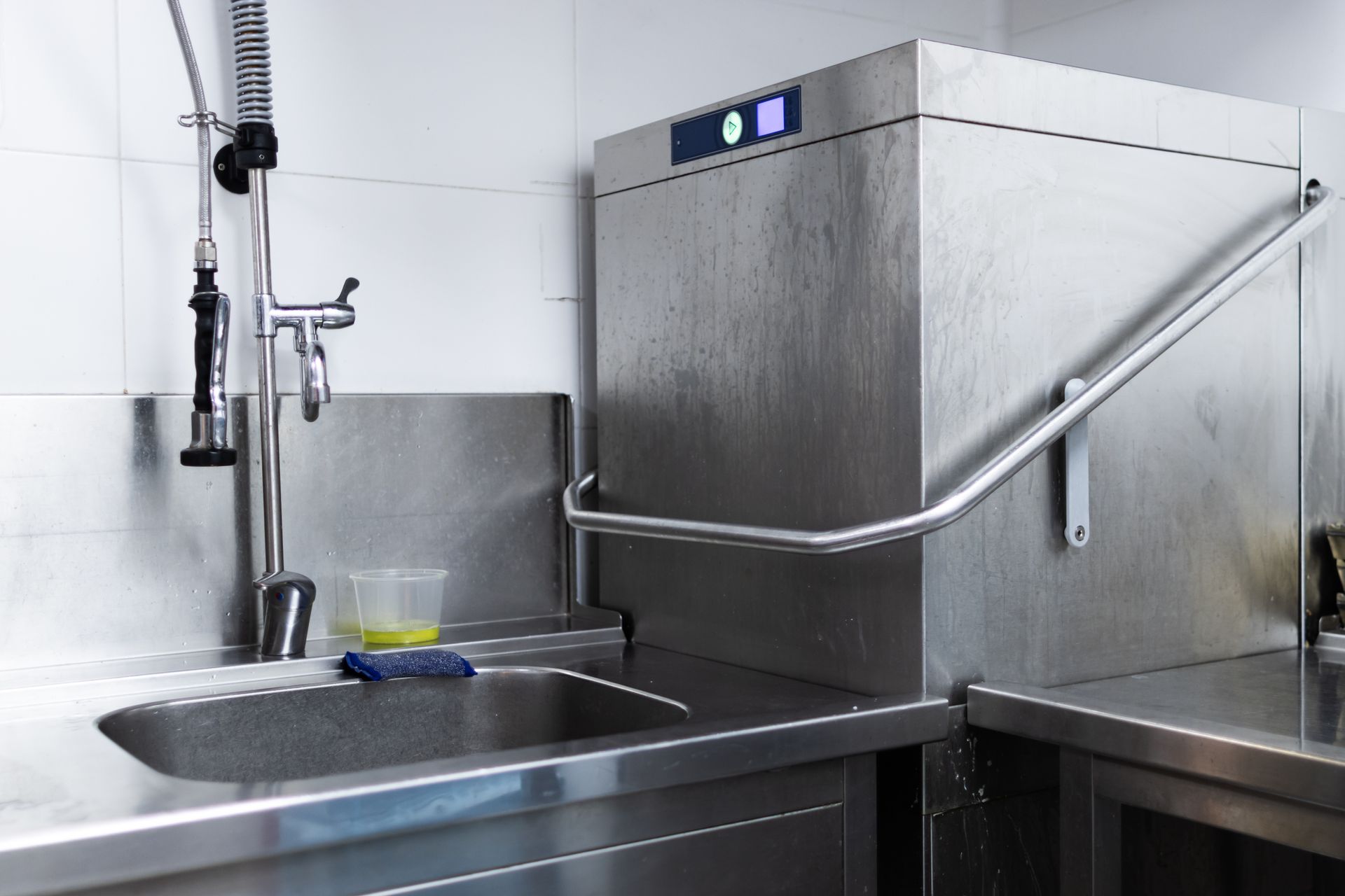 Un lave-vaisselle commercial en acier inoxydable et un évier avec robinet à douchette dans une cuisine professionnelle.