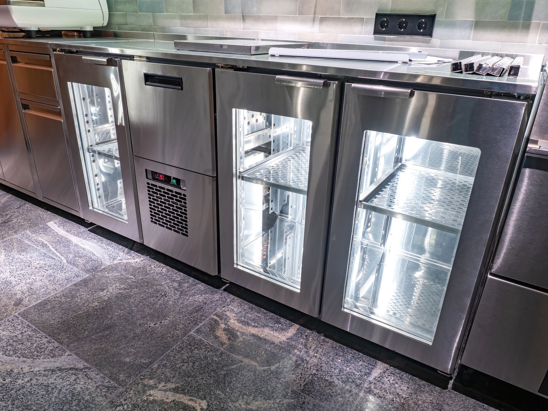 Une rangée de réfrigérateurs commerciaux sous comptoir en acier inoxydable, avec portes vitrées et éclairage intérieur, sur un sol en pierre.