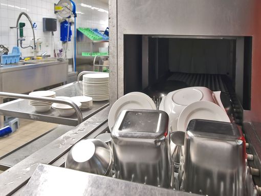 Dans une cuisine professionnelle, des récipients et des assiettes en acier inoxydable reposent sur le plateau d'un lave-vaisselle industriel.