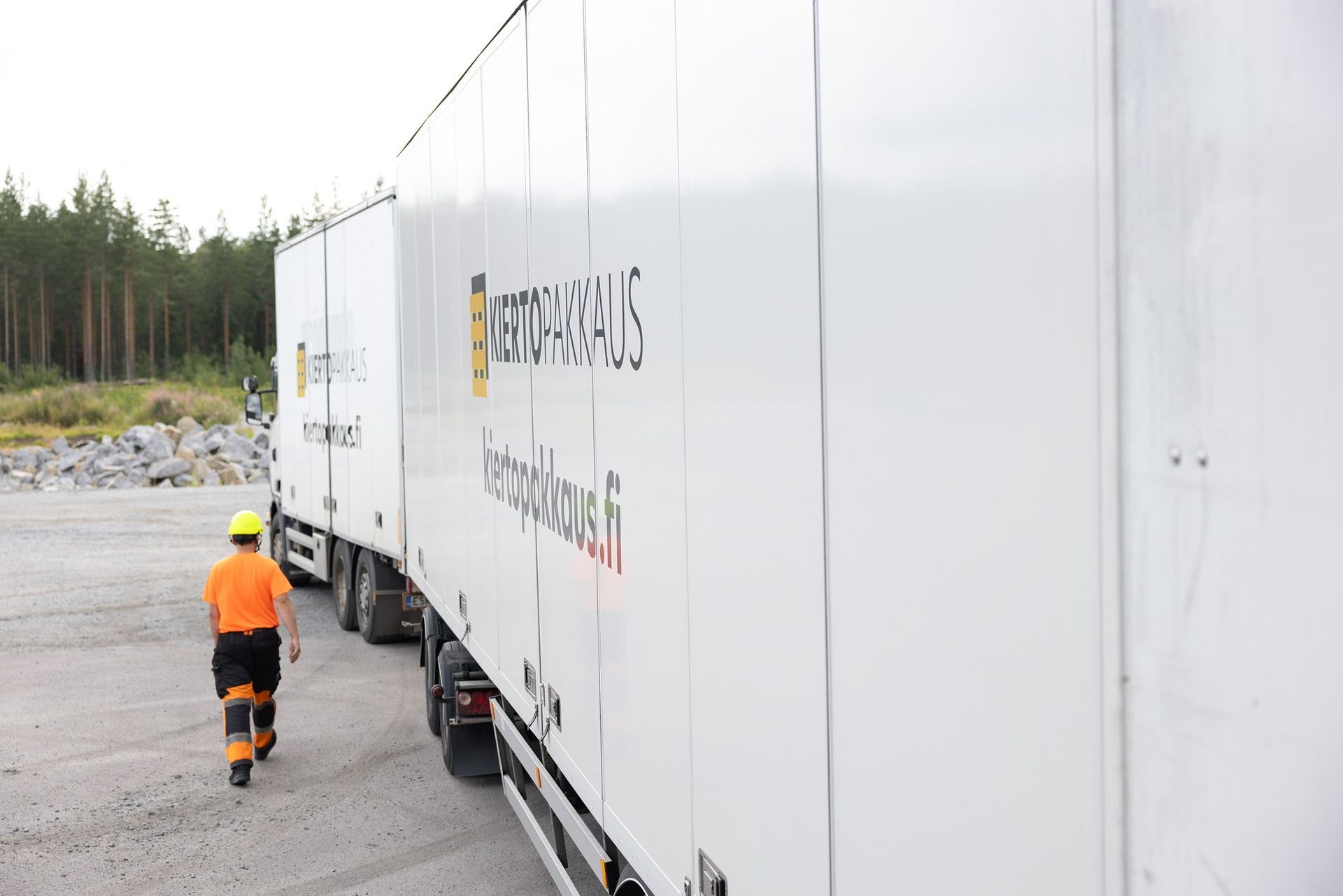 Heijastinoranssiin vaatetukseen pukeutunut työntekijä kävelee sorakentälle pysäköidyn valkoisen puoliperävaunun vierellä.