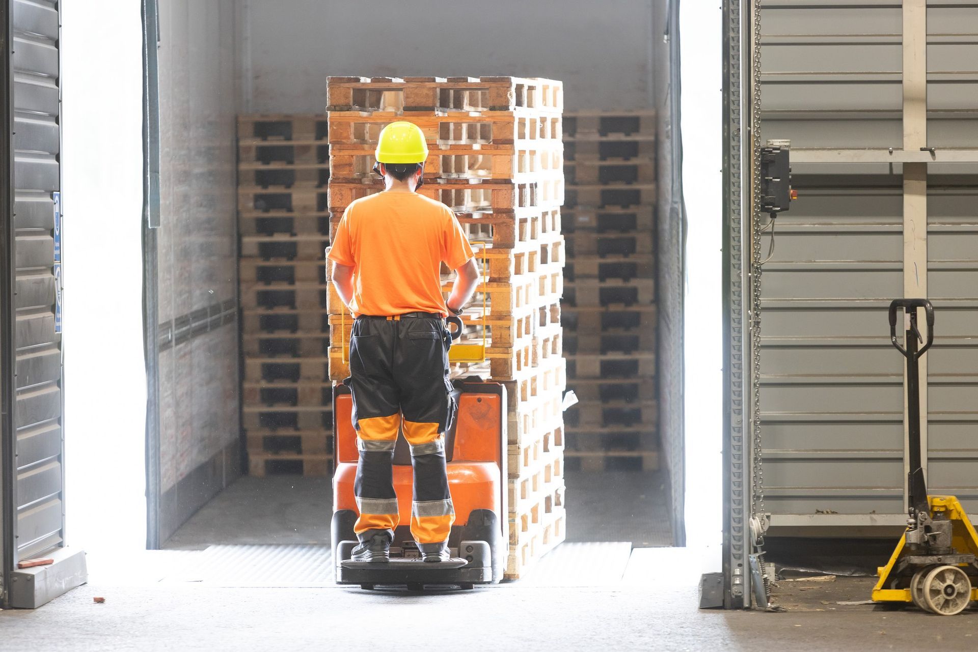 Keltaiseen kypärään ja oranssiin paitaan pukeutunut työntekijä käyttää sähkökäyttöistä lavanosturia ja kuljettaa lavapinoa.