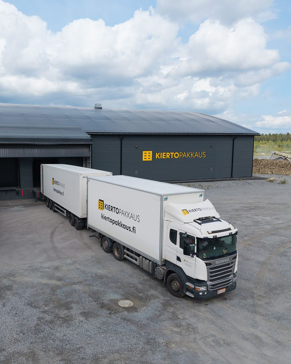 Valkoinen puoliperävaunullinen rekka pysäköitynä tumman, keltaisella logolla varustetun varastorakennuksen eteen pilvisen taivaan alla.