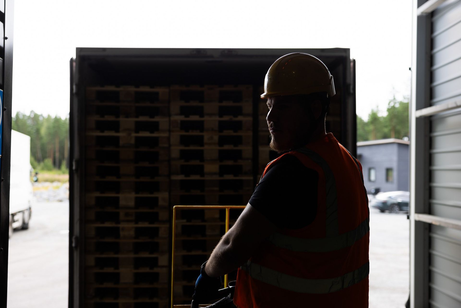 Heijastinliiviin ja kypärään pukeutunut työntekijä seisoo pinottujen puulavojen täyttämän perävaunun edessä.