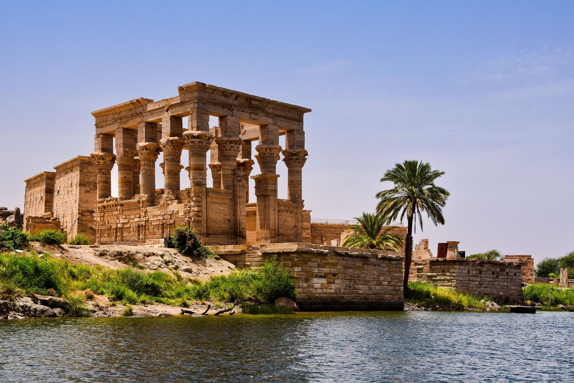 Ancient stone temple ruins by a river, with a palm tree and blue sky