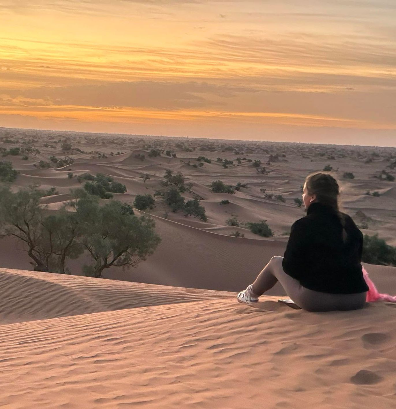Woman seated on a sand dune, looking at a desert sunset.