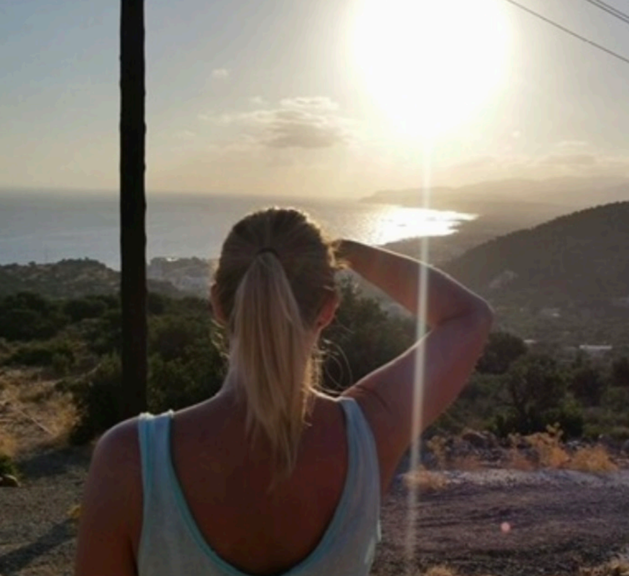 Woman with blonde ponytail looking at sunset over ocean and hills.