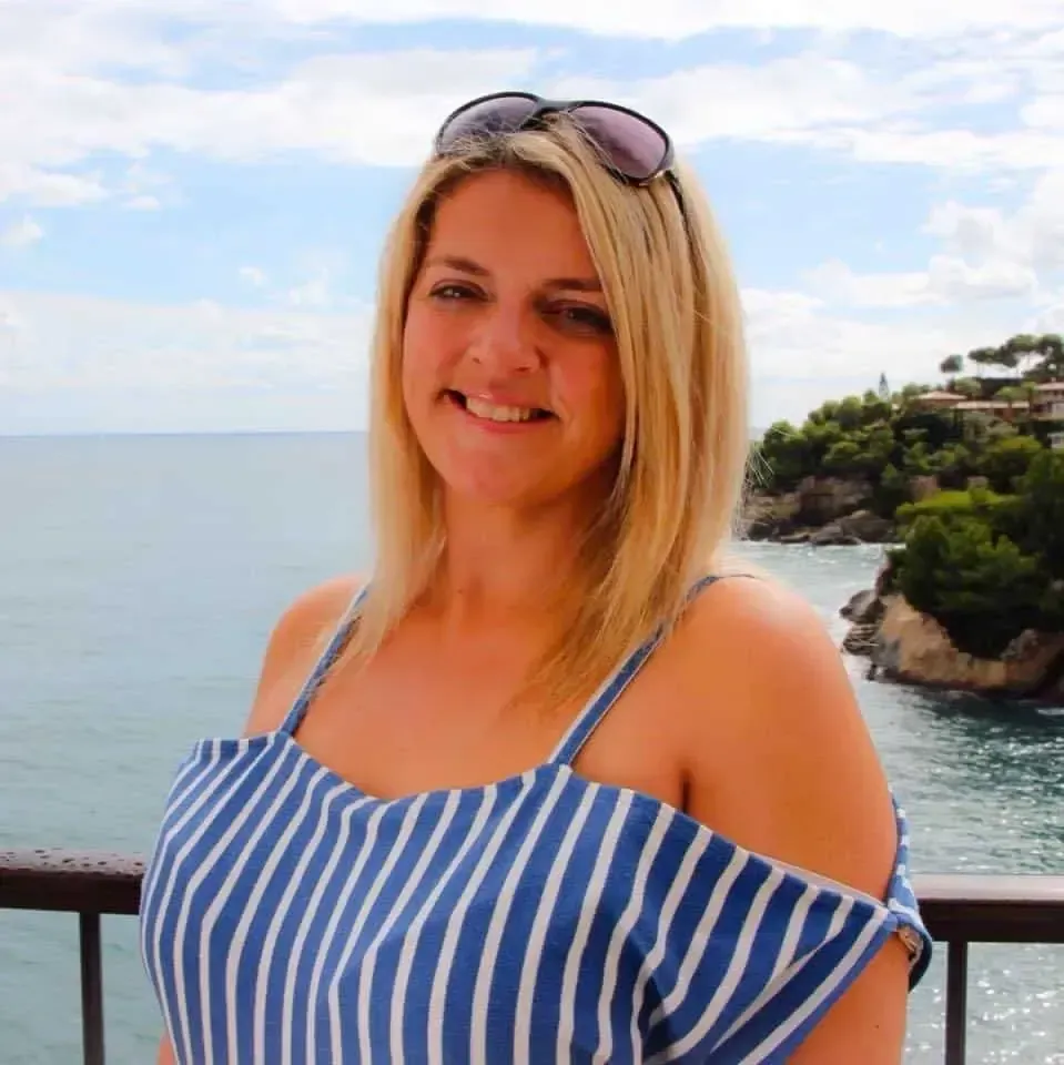Woman with blonde hair wearing a striped top, smiling near a body of water with a cloudy sky.