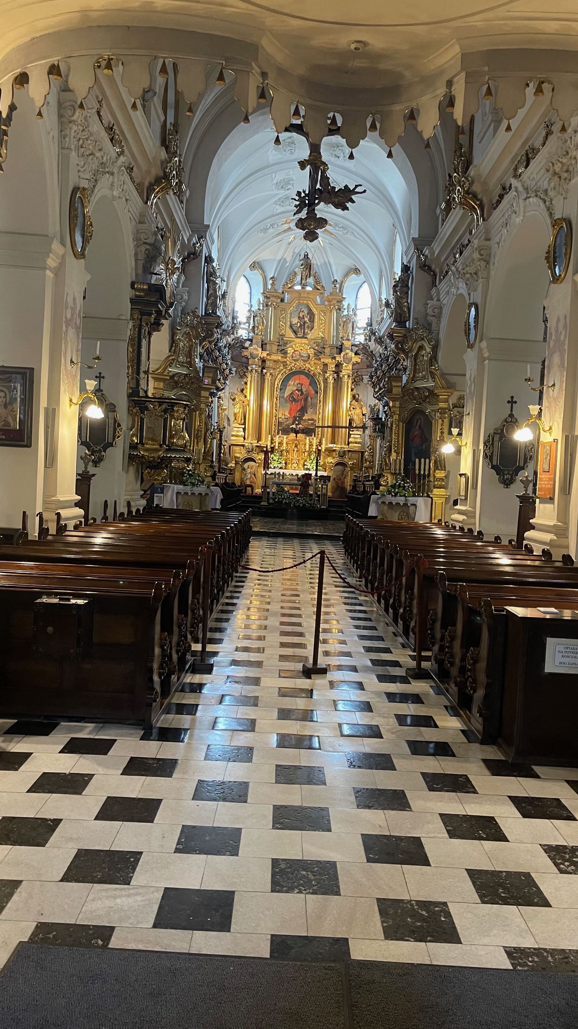 Interior of a church with ornate gold altar, rows of wooden pews, and checkered floor.