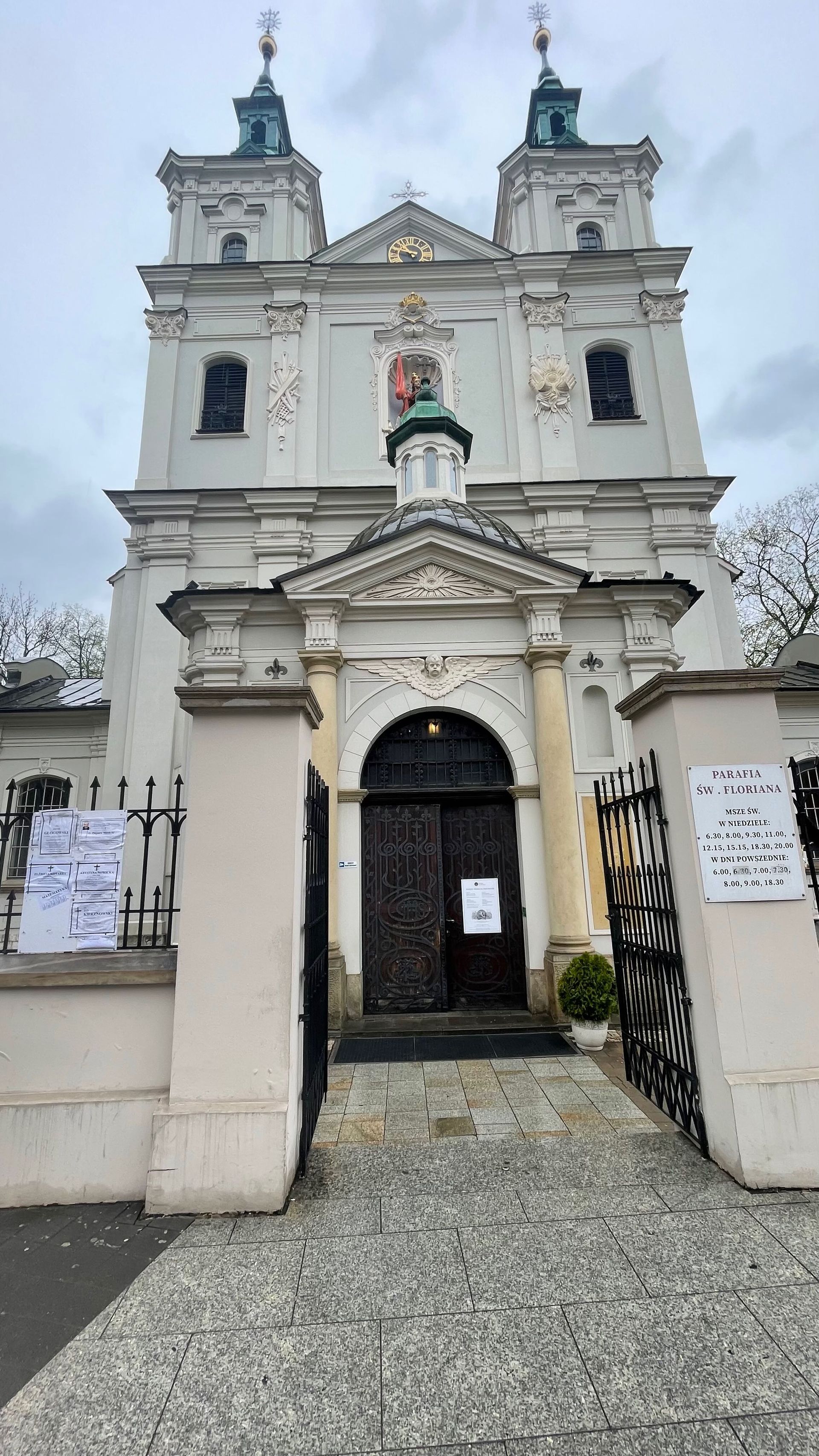 White church with two towers, entrance, dark doors, and gate. Paved walkway.