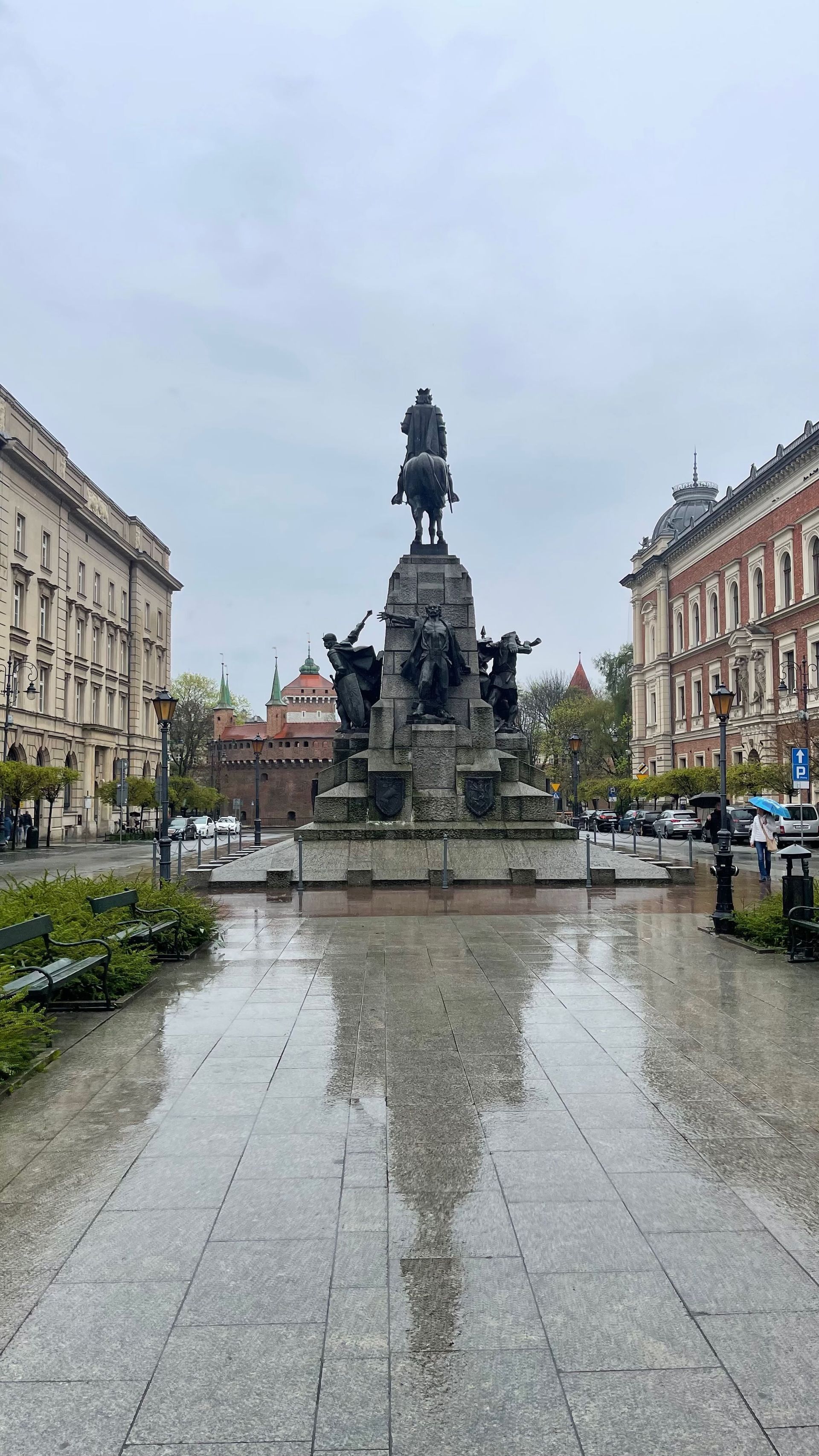 Monument on a wet cobblestone street, flanked by buildings. Cloudy sky.