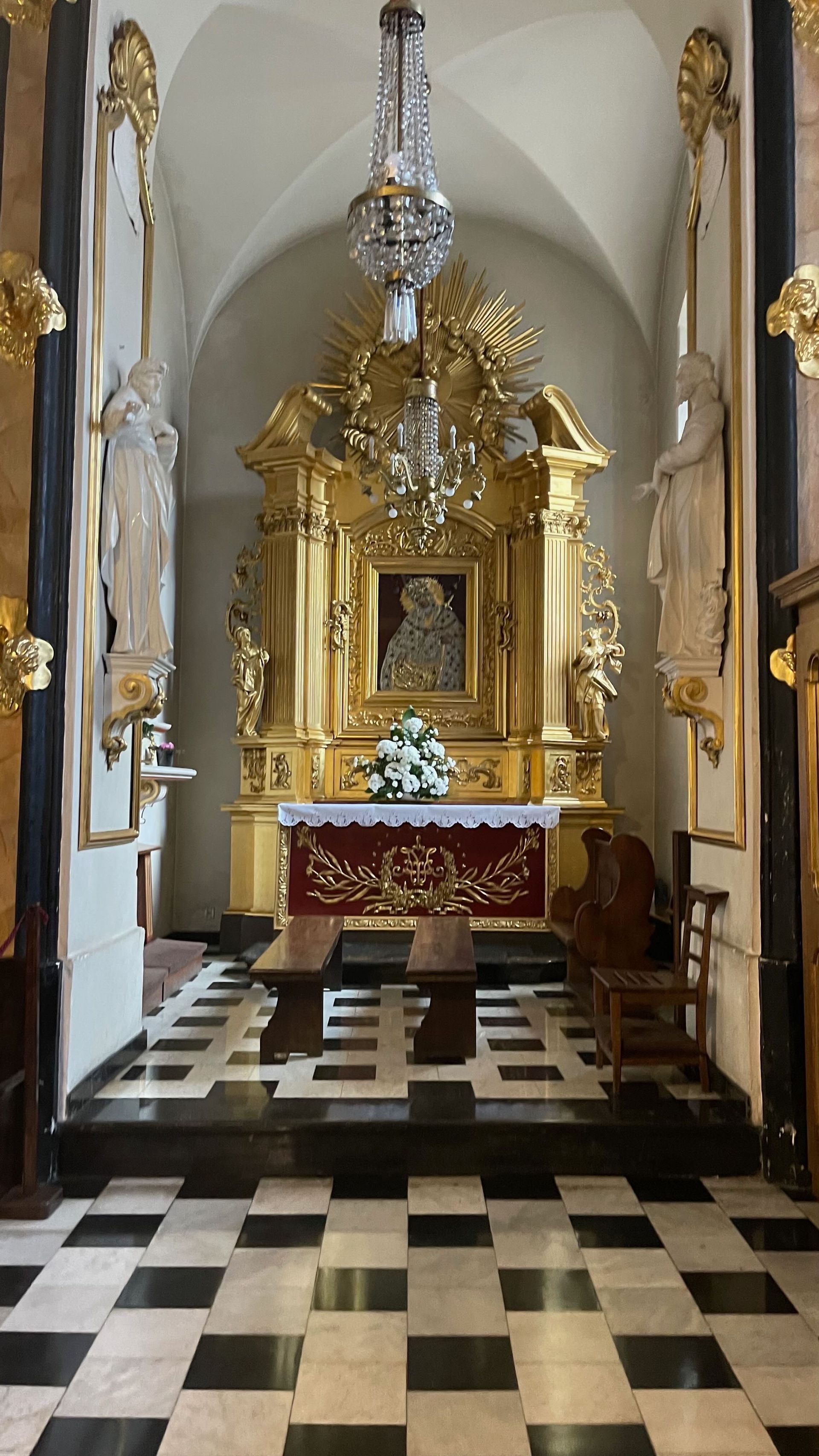 Ornate gold altar in a church, featuring a painting, flowers, and chandelier. Black and white tiled floor.