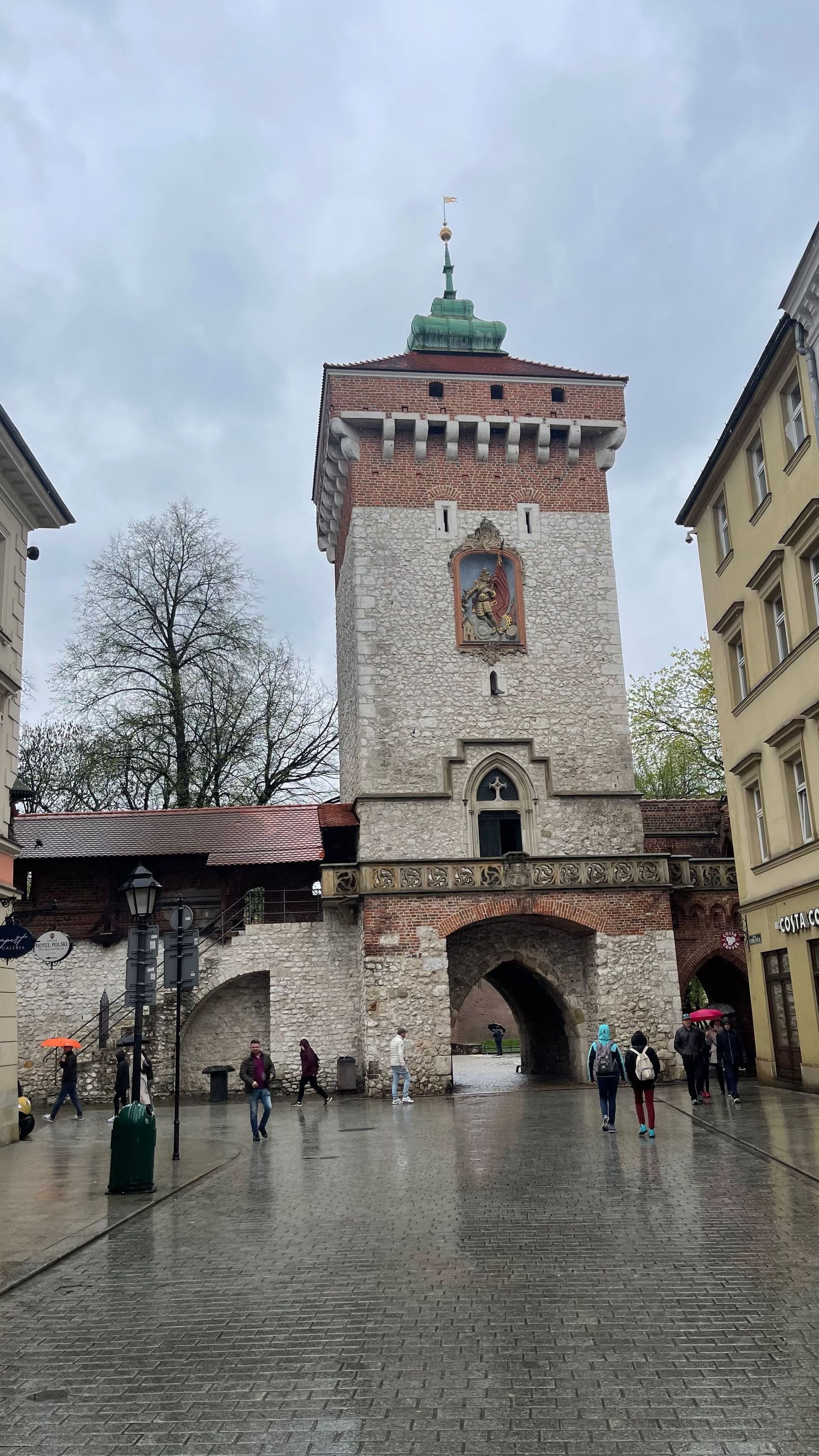 Medieval brick tower with arched gateway in a wet, cobblestone square; people walking.