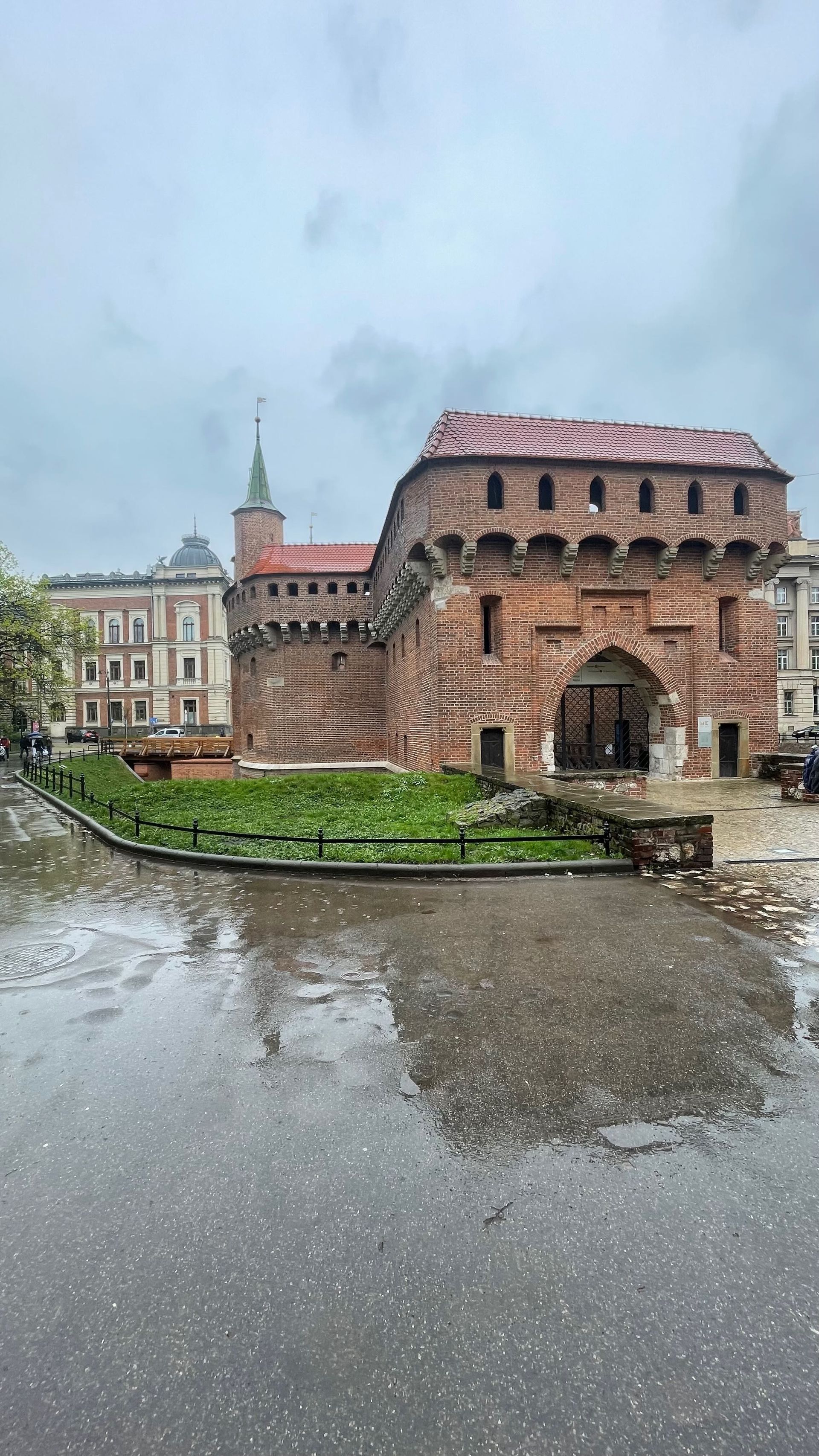 Brick Barbican building and adjacent structures in Krakow, Poland, reflecting in wet pavement under a cloudy sky.