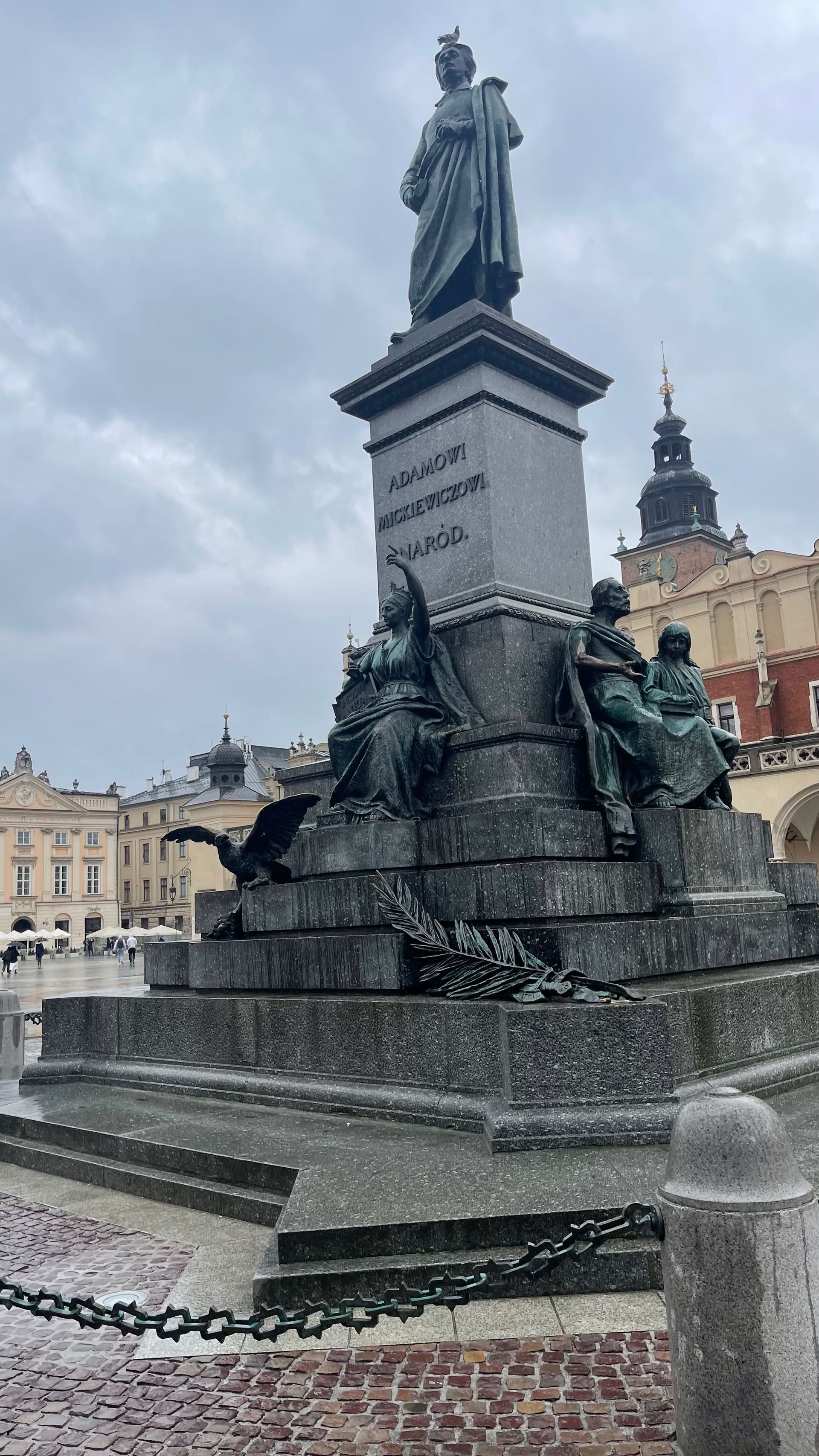 Monument to Adam Mickiewicz in Krakow's Main Market Square. Bronze statue atop a tiered base, cloudy sky.