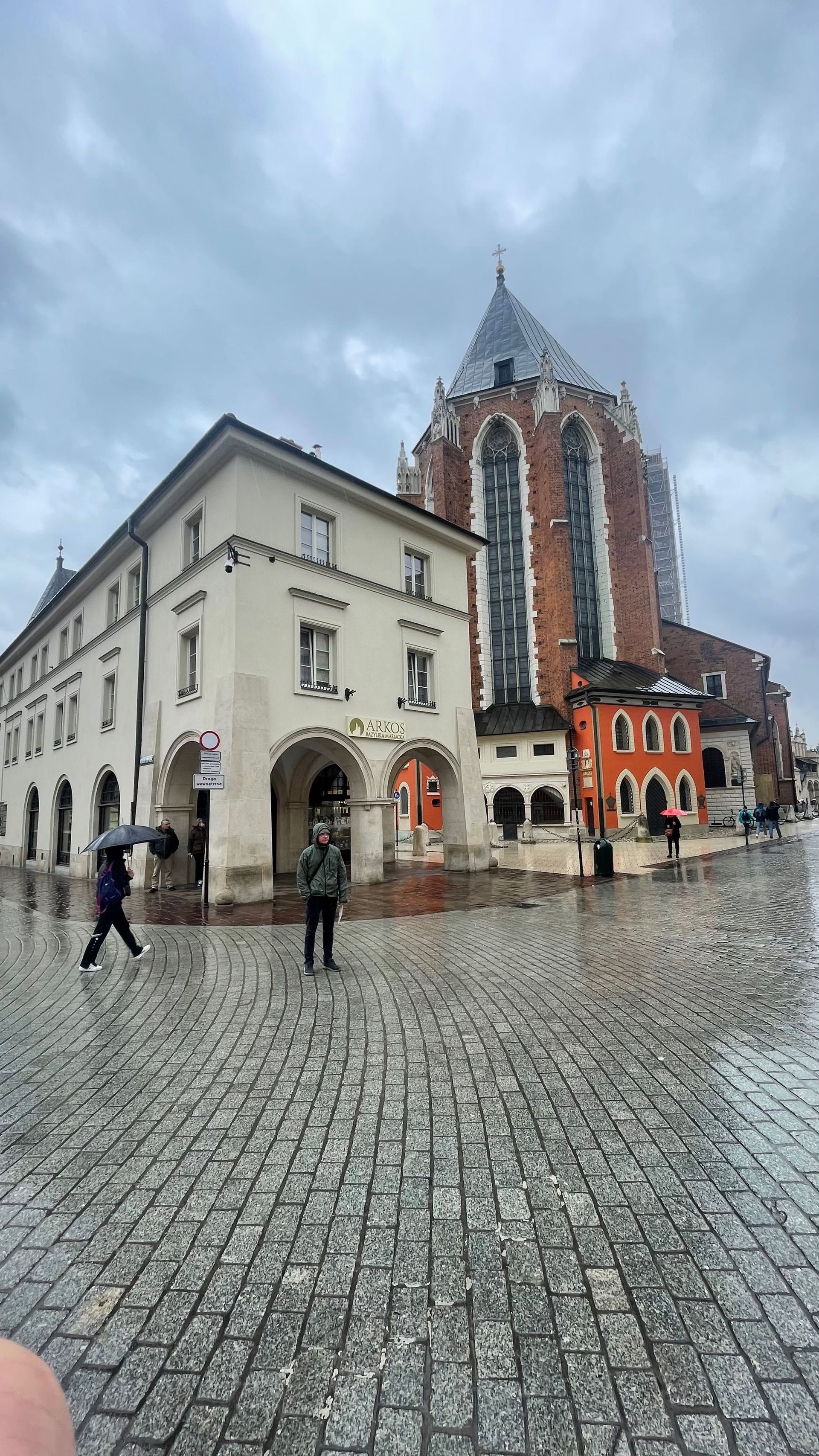 A rainy day in Kraków, Poland. People walk near the historic town hall and St. Mary's Basilica on a cobblestone street.