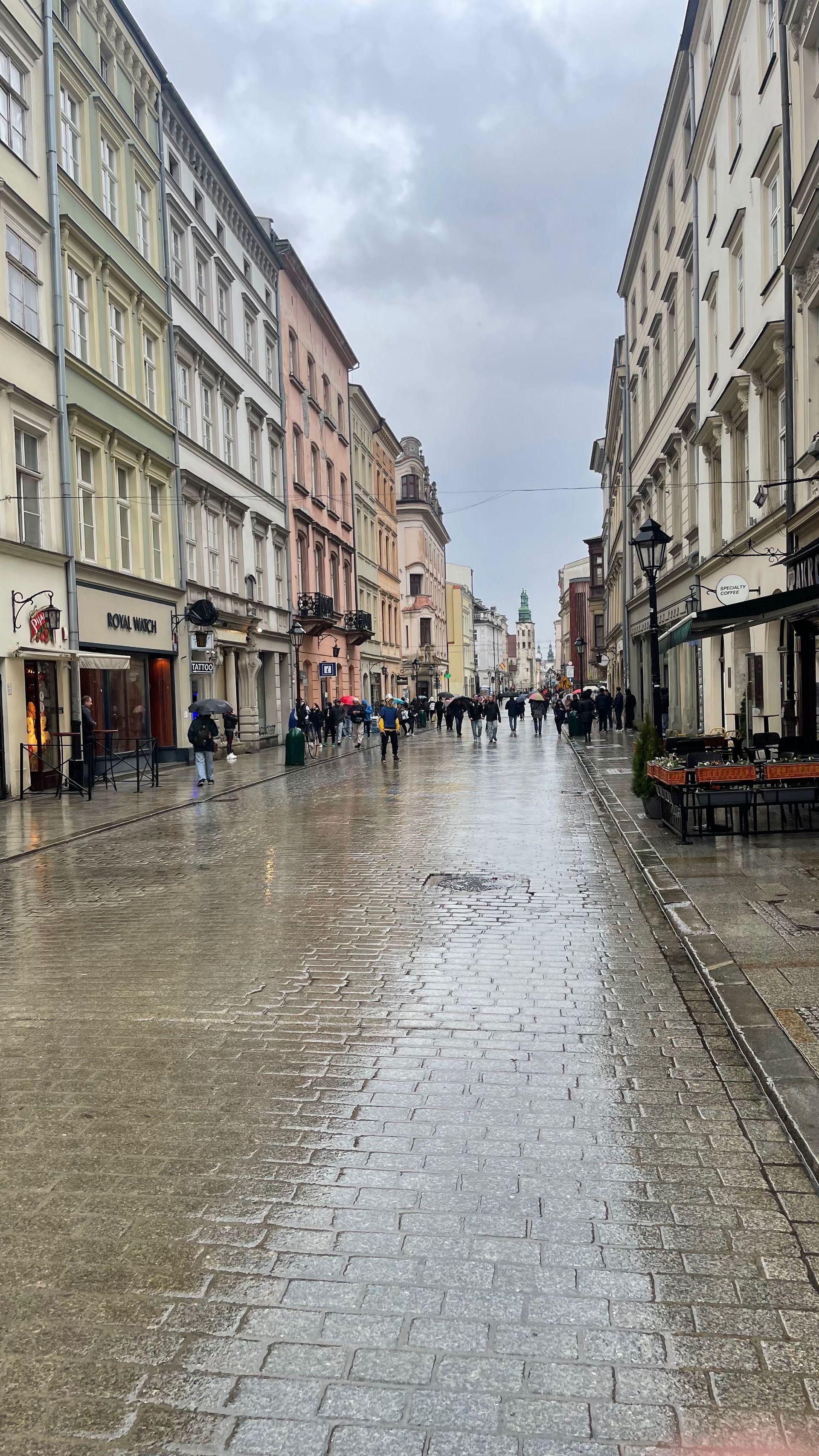 Wet cobblestone street lined with buildings; pedestrians walk under overcast sky.
