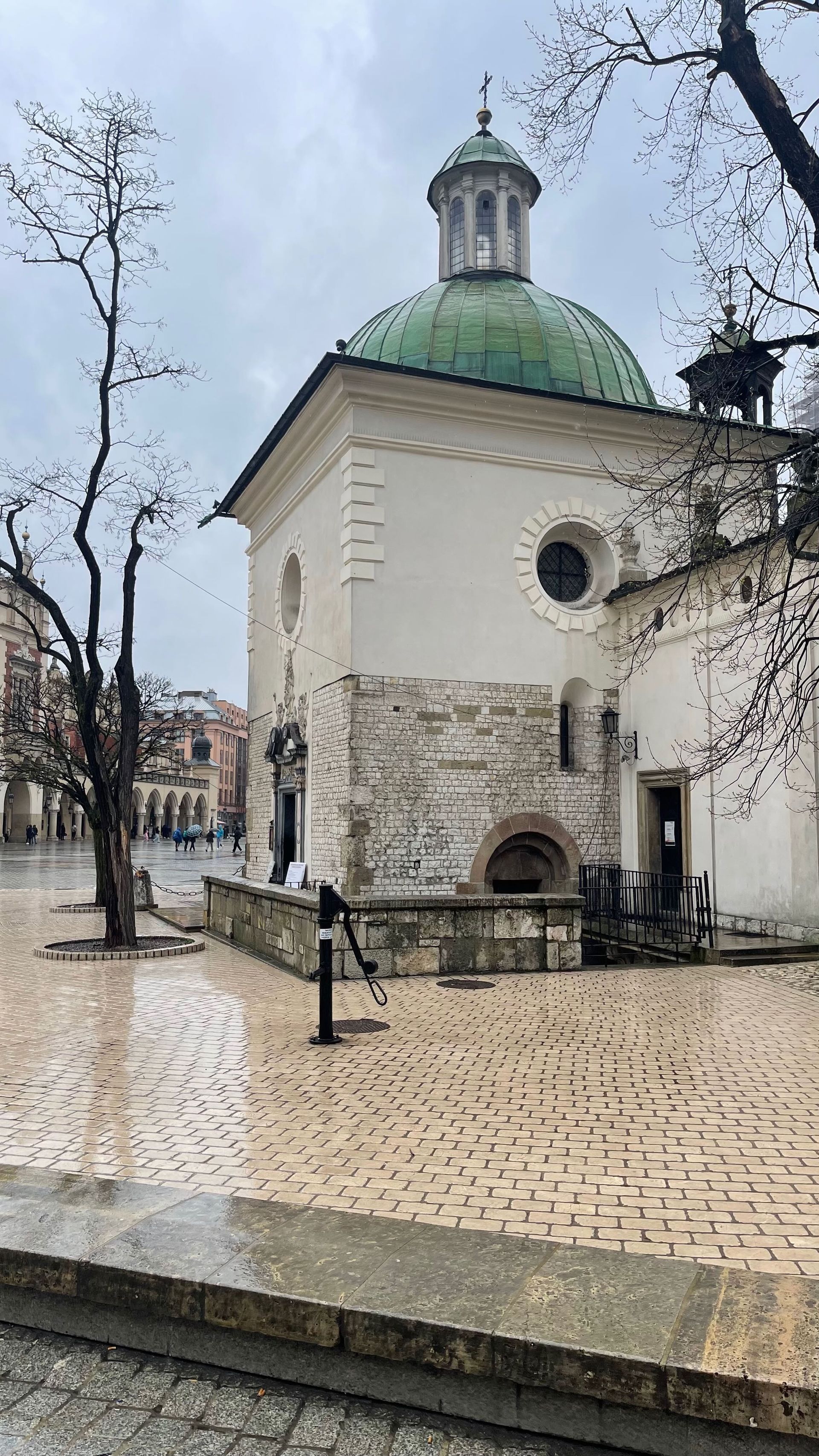 White building with green dome, trees, and cobblestone square on a cloudy day.