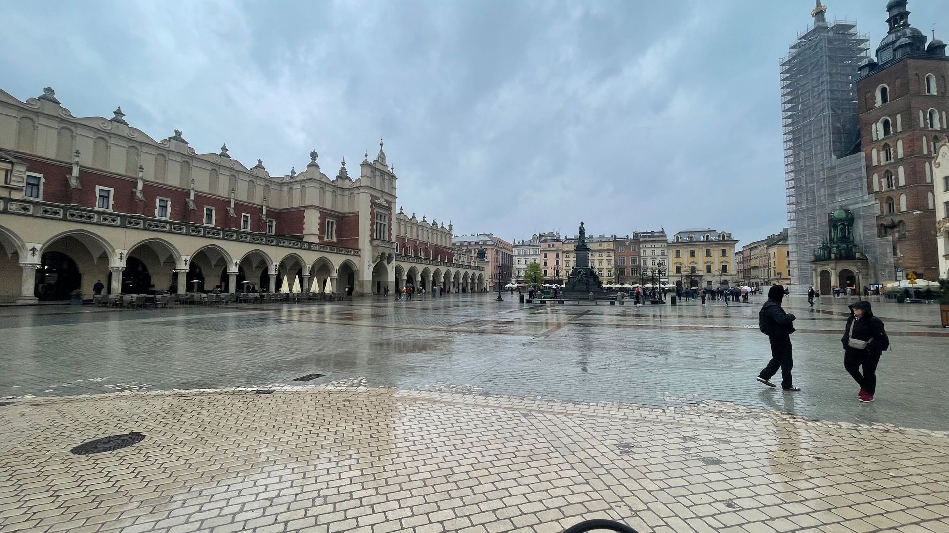 Main Square in Krakow, Poland, with historic buildings, reflecting wet cobblestones, and two people walking.