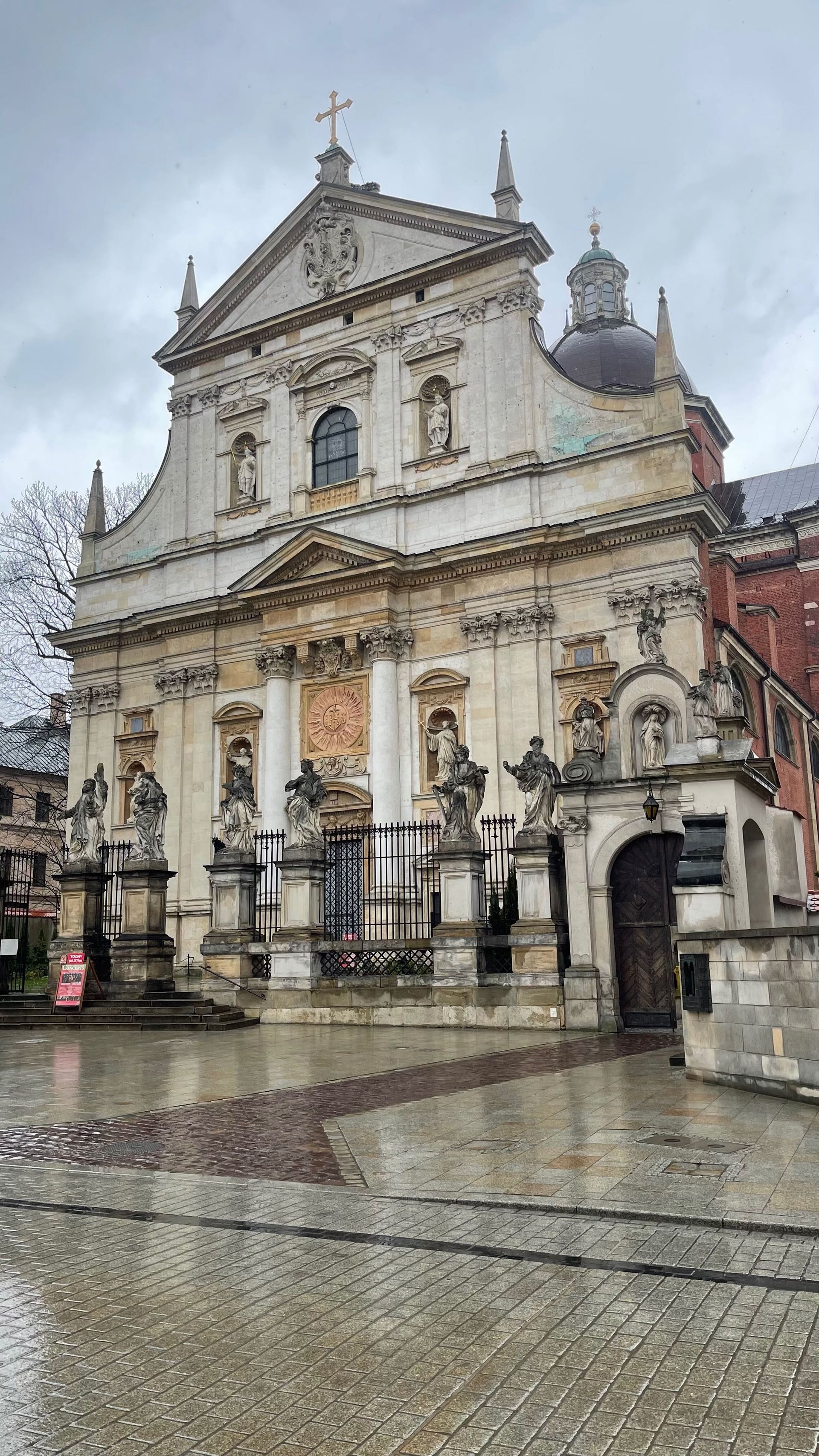 Baroque-style church facade with statues, ornate details, and a dark wooden door on a wet cobblestone square.