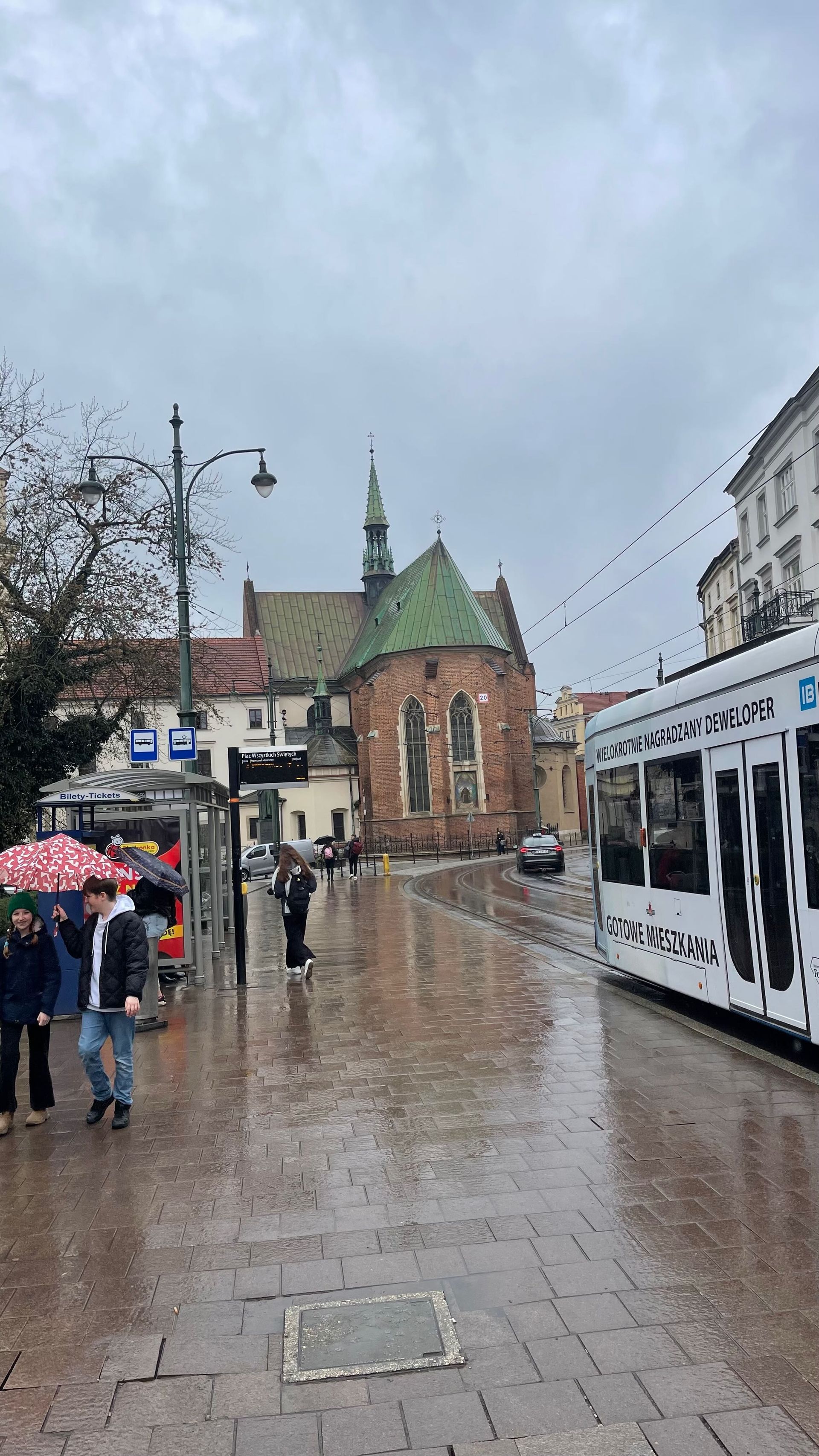 Street scene in rain, featuring a church, tram, and people with umbrellas. Wet pavement reflects the overcast sky.