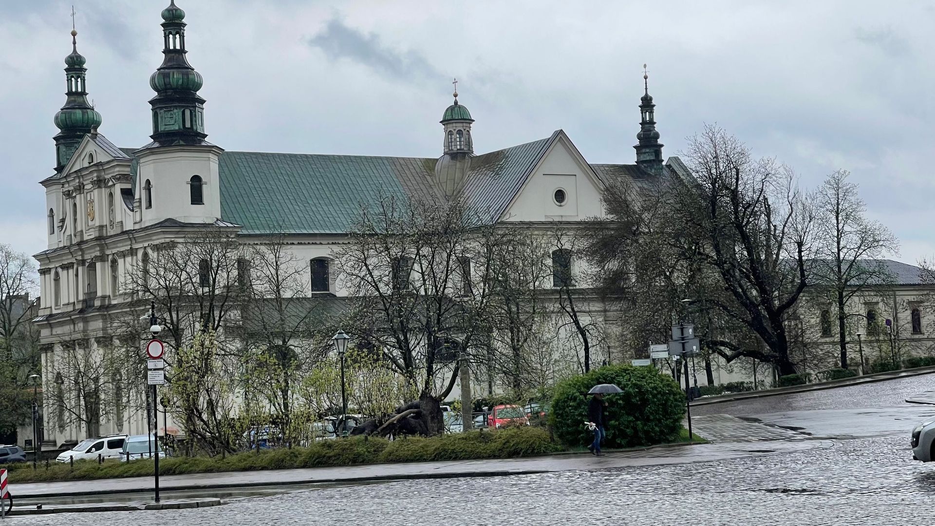 White church with green roof and several spires under a cloudy sky. Trees and a wet street in front.