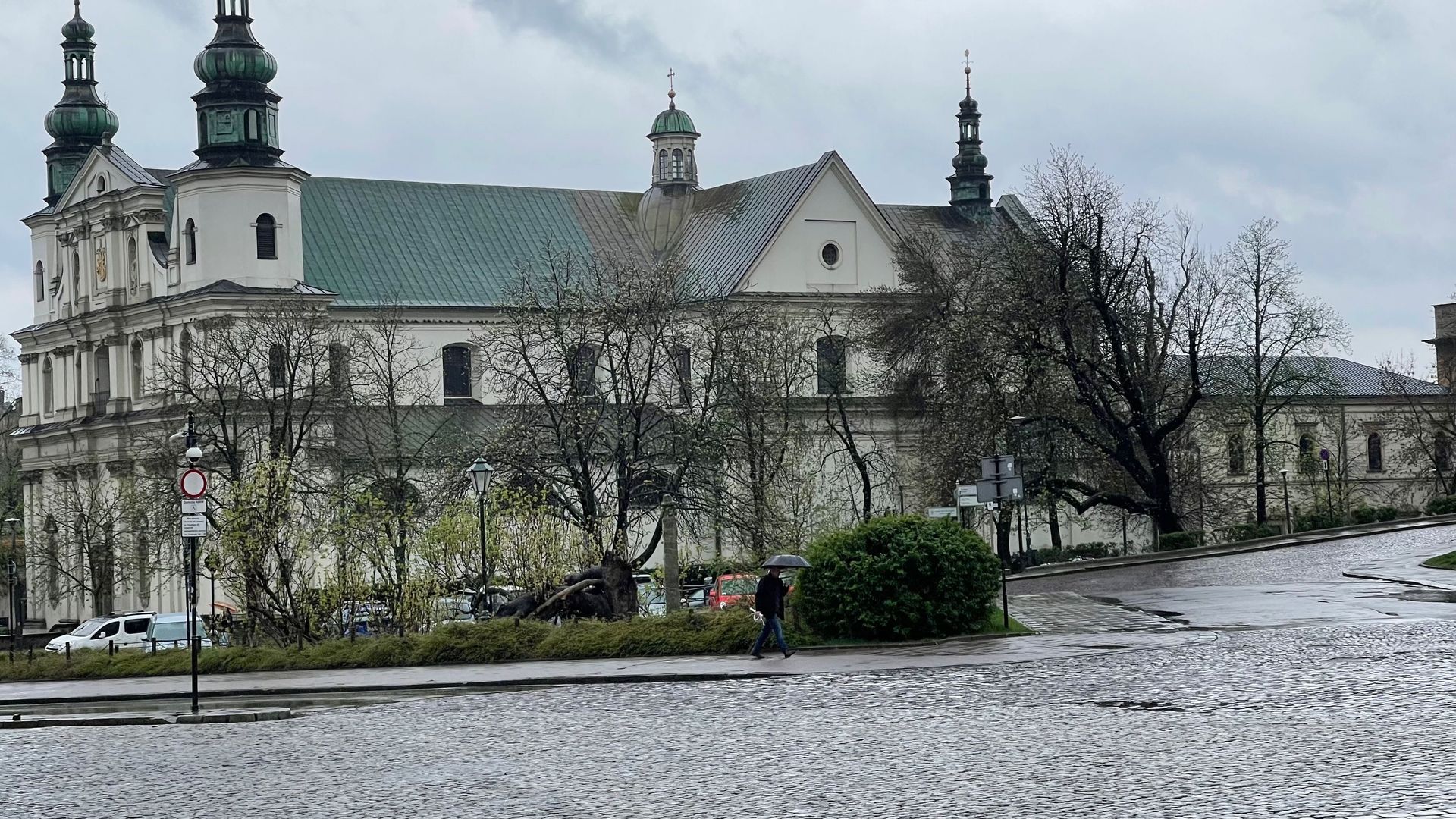 Stone church with green and white towers, trees, and overcast sky. A person walks on cobblestone street.