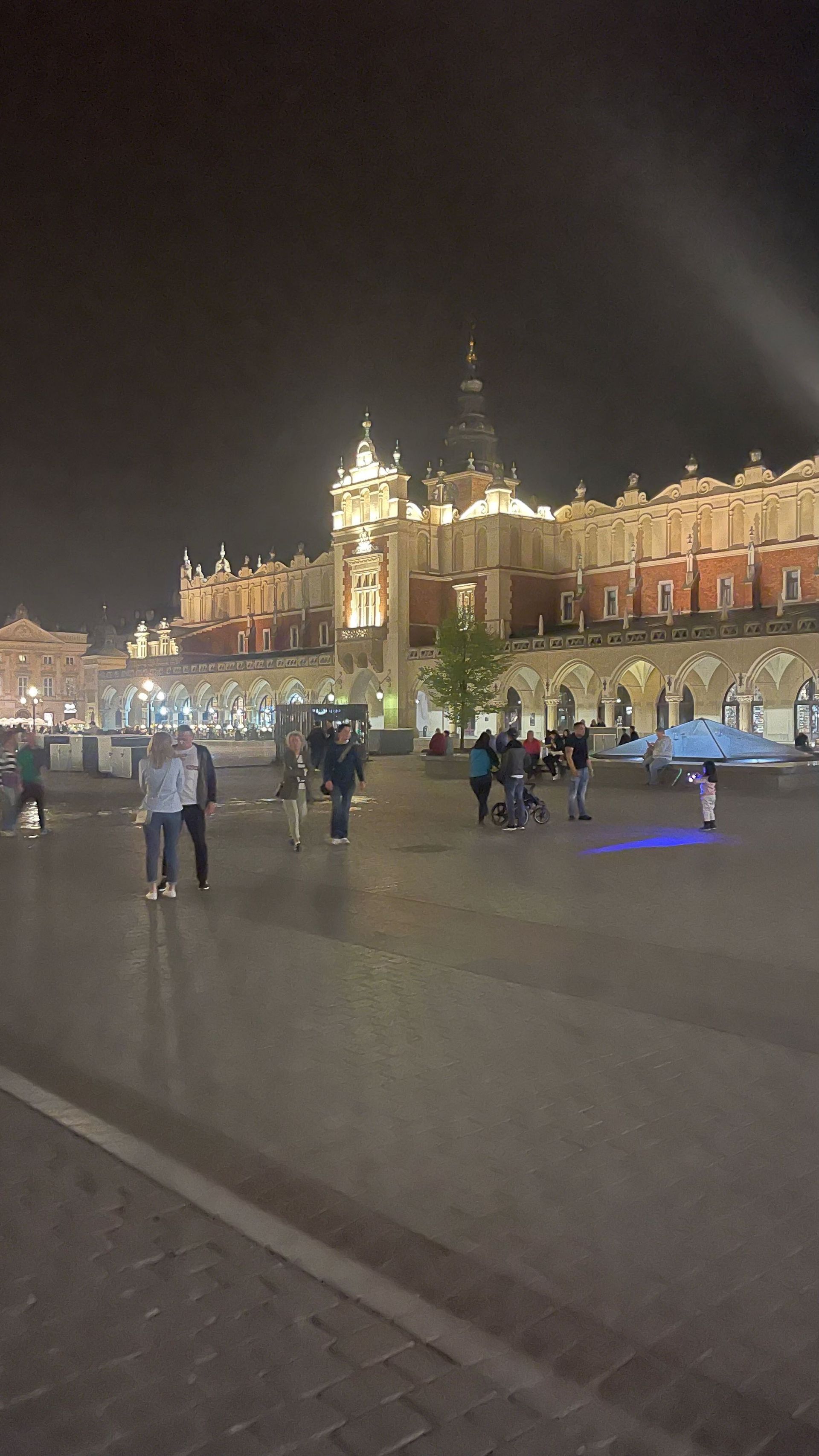 Nighttime view of a historic building in a town square, illuminated with lights, with people walking around.