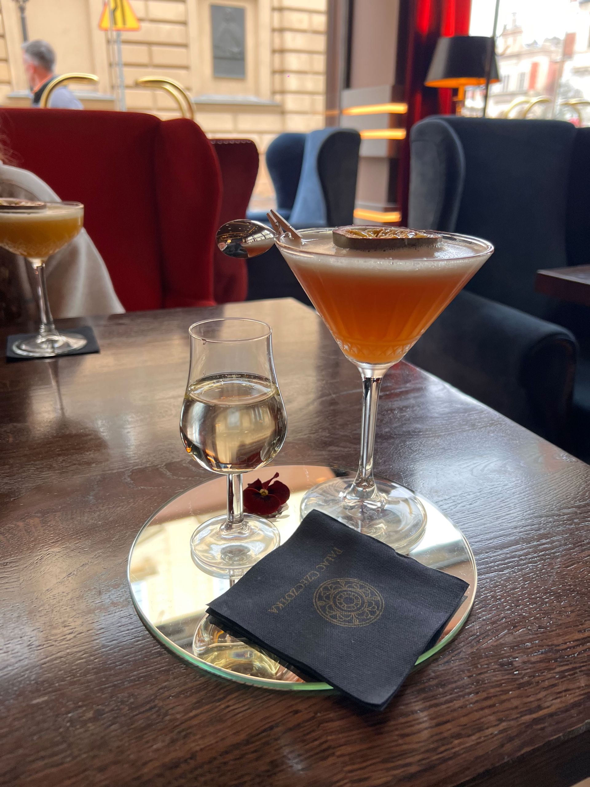 Cocktail with passion fruit garnish, shot glass, and black napkin on a mirrored coaster, set on a dark wood table.