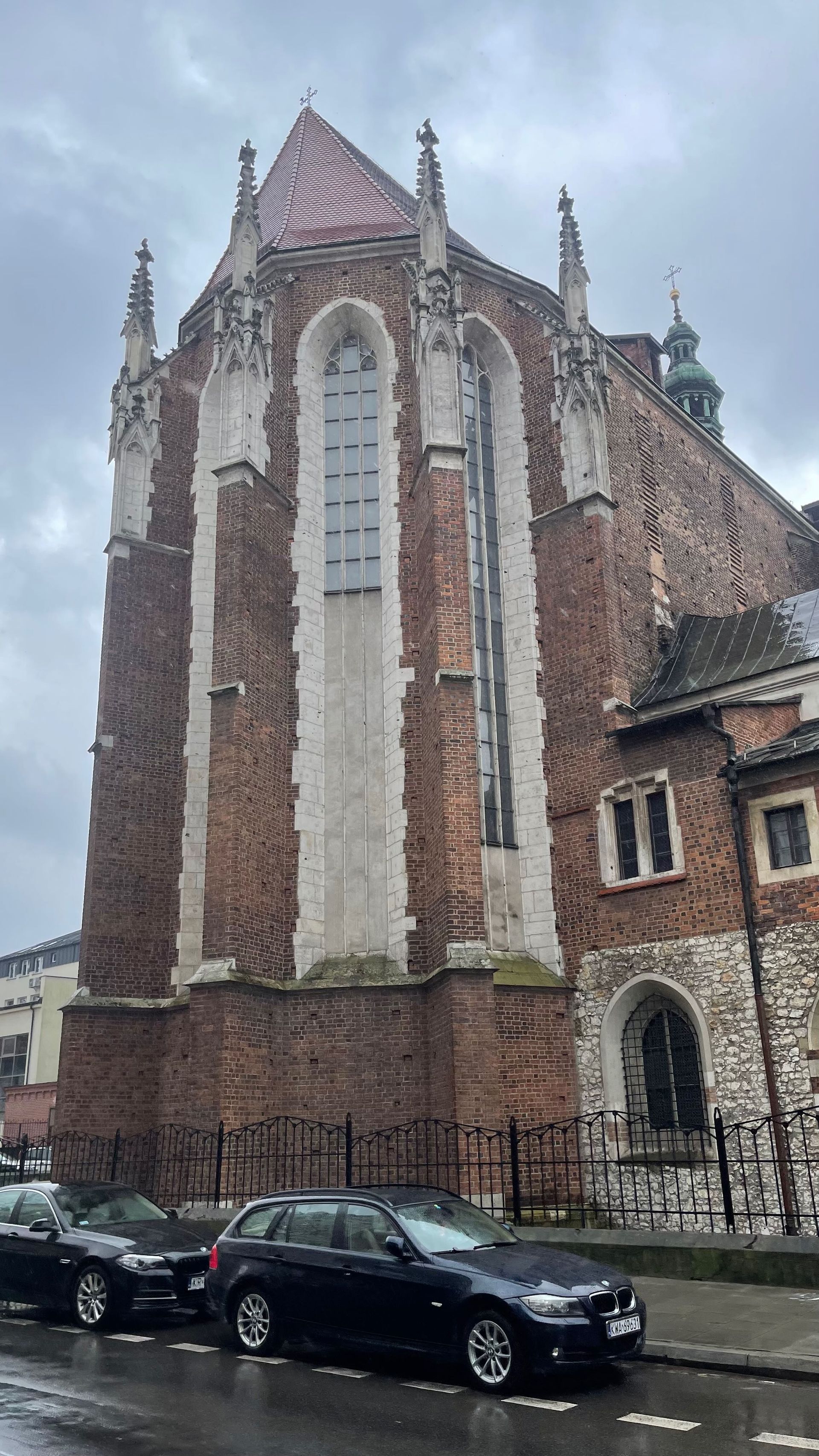 Side view of a tall, Gothic brick building with a patterned red tile roof, cars parked in front.