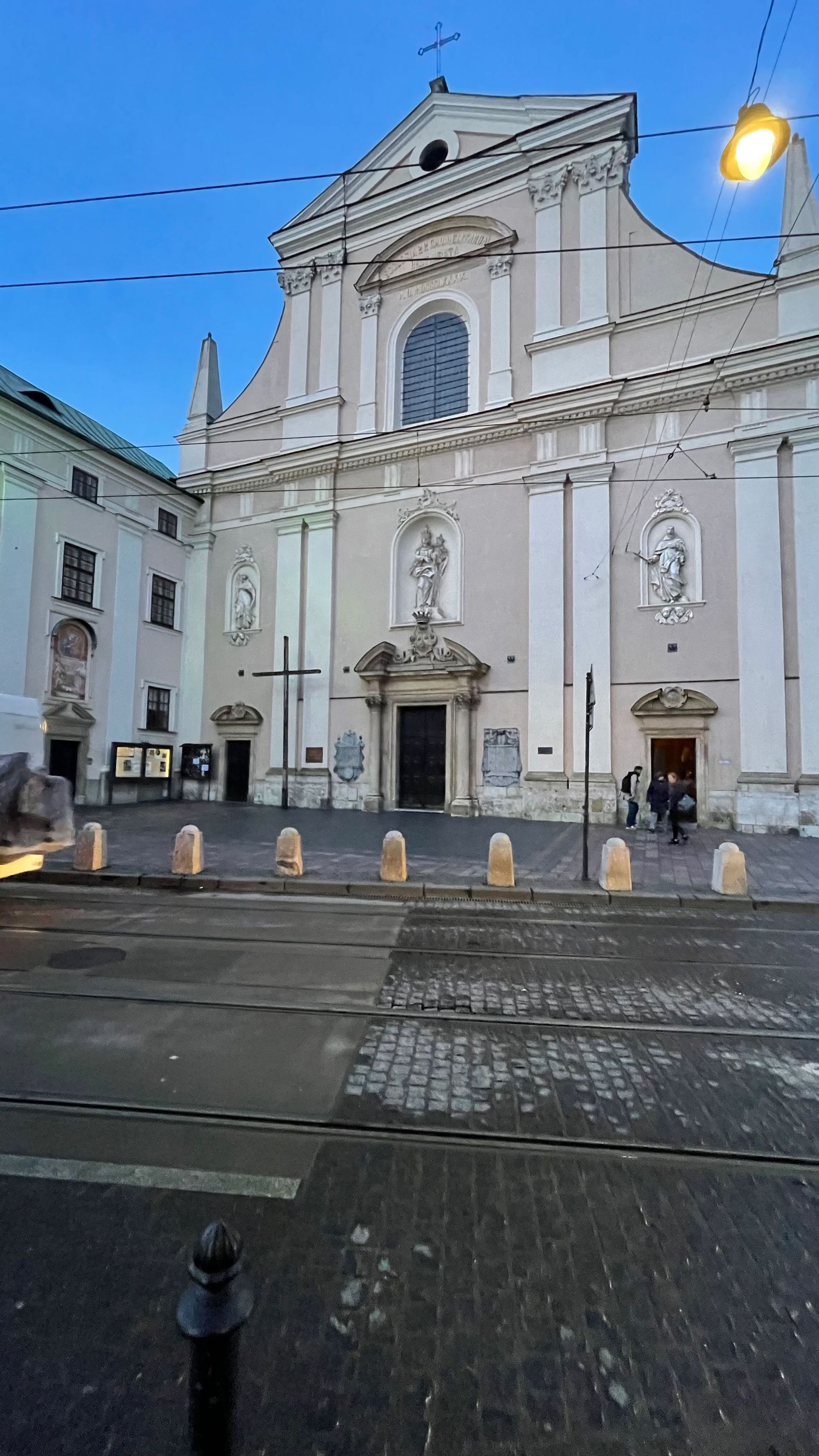 Baroque church exterior with light beige facade, cross, and cobblestone plaza.