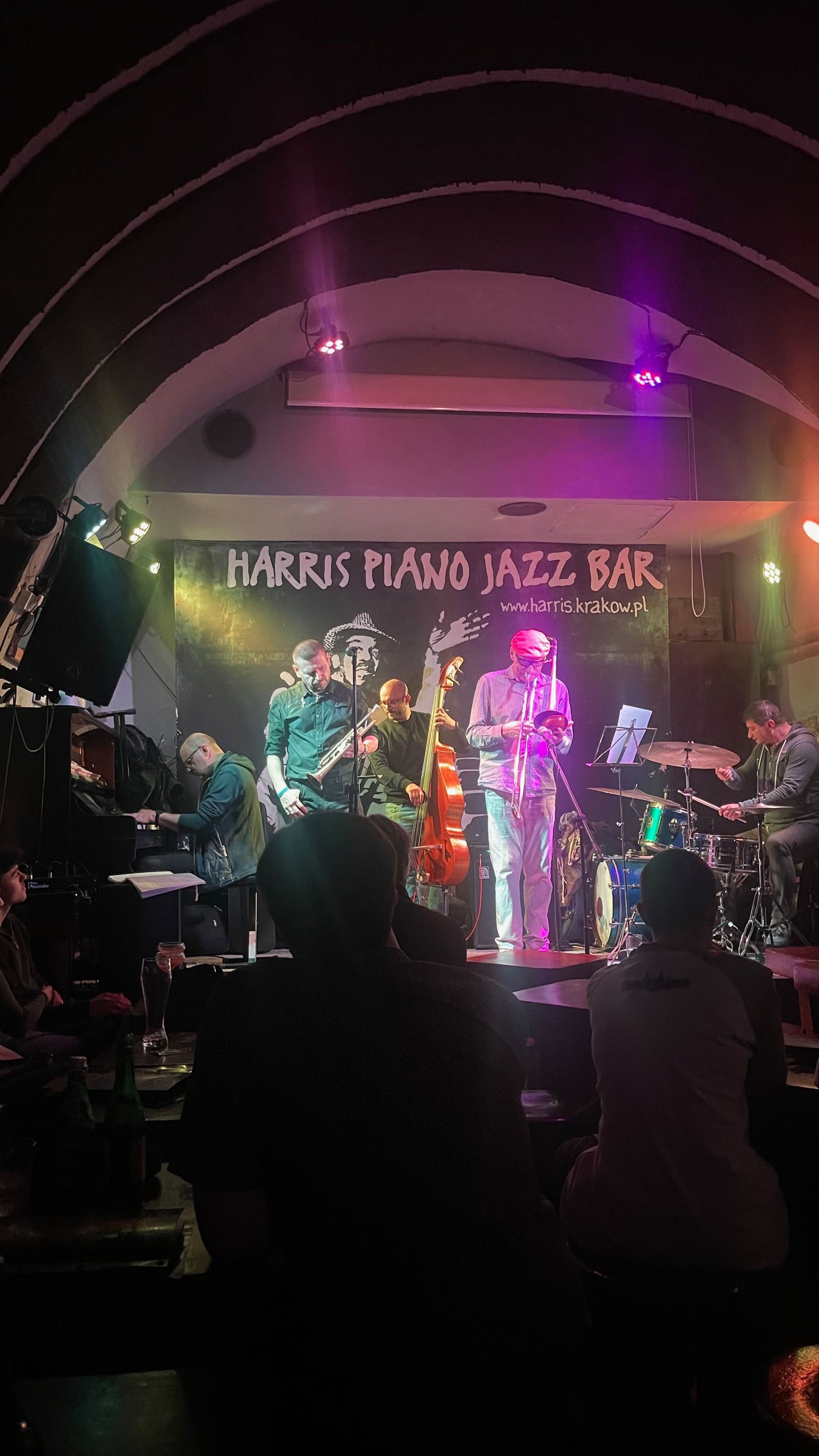 Jazz band performing at Harris Piano Jazz Bar. Stage lit with colorful lights. Audience seated in front.