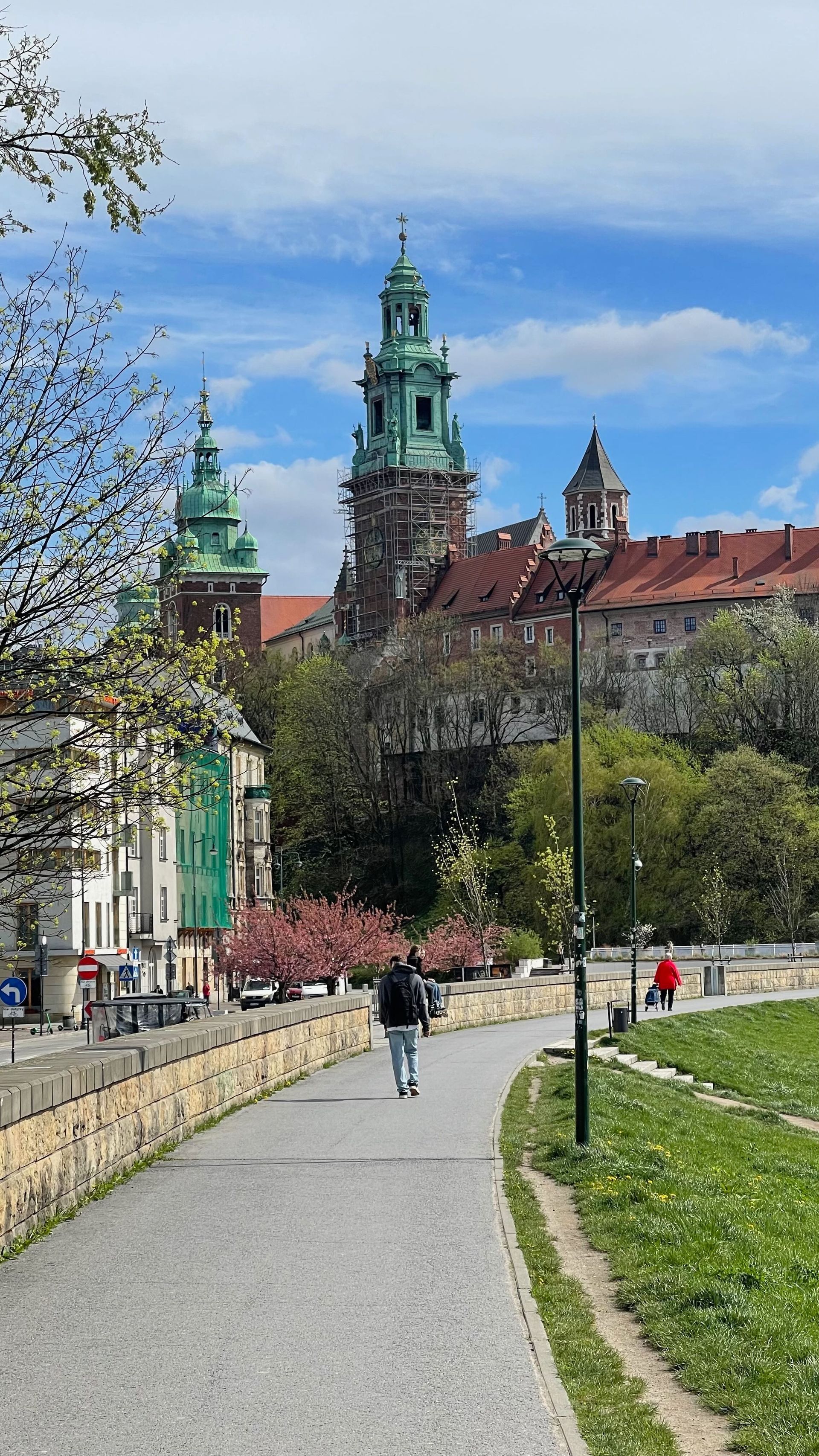 Person walks along a path towards Wawel Castle in Kraków, Poland. Green towers and blue sky in background.