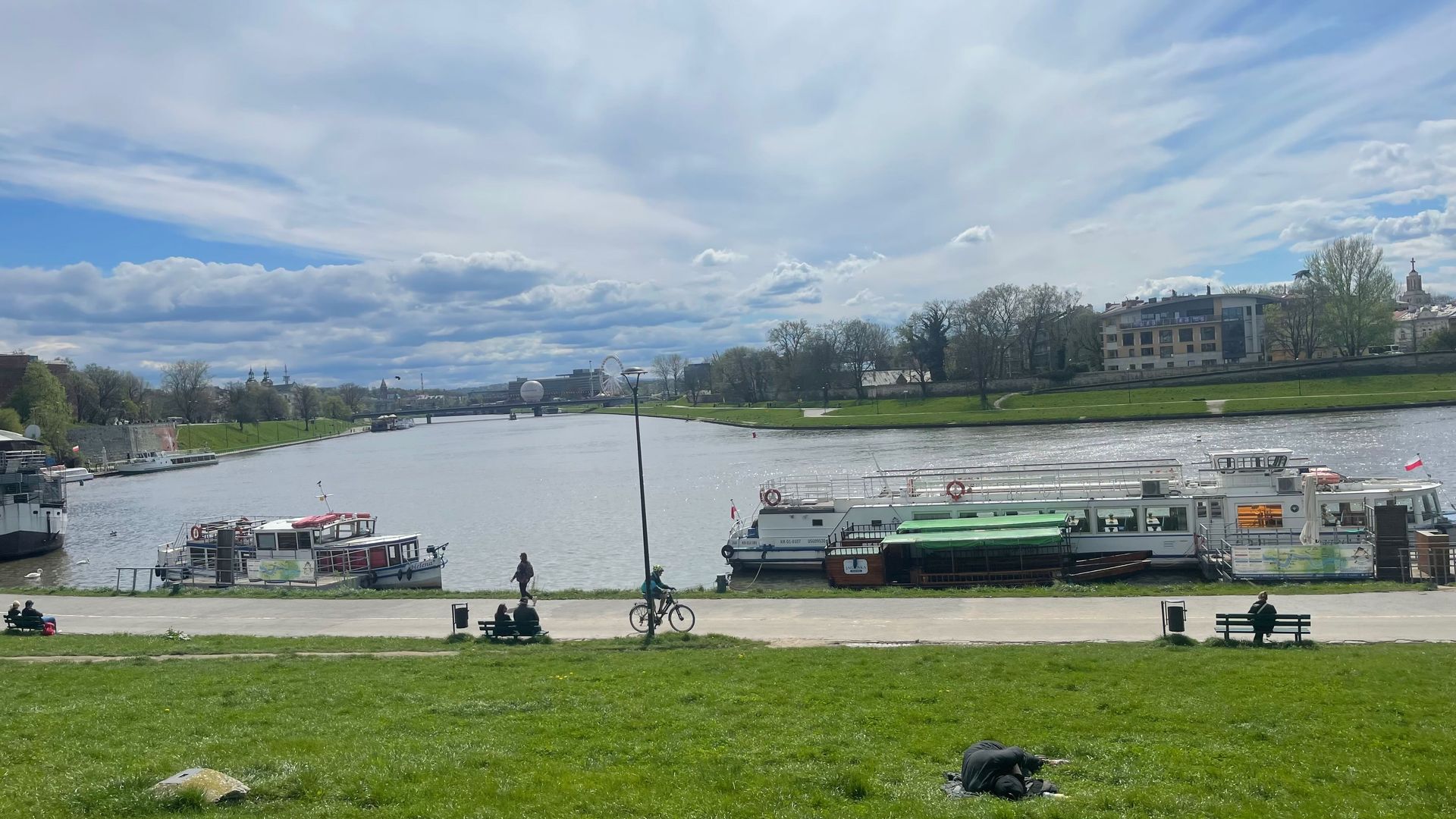 River with boats, park, and cloudy sky. People are relaxing on benches near the water.